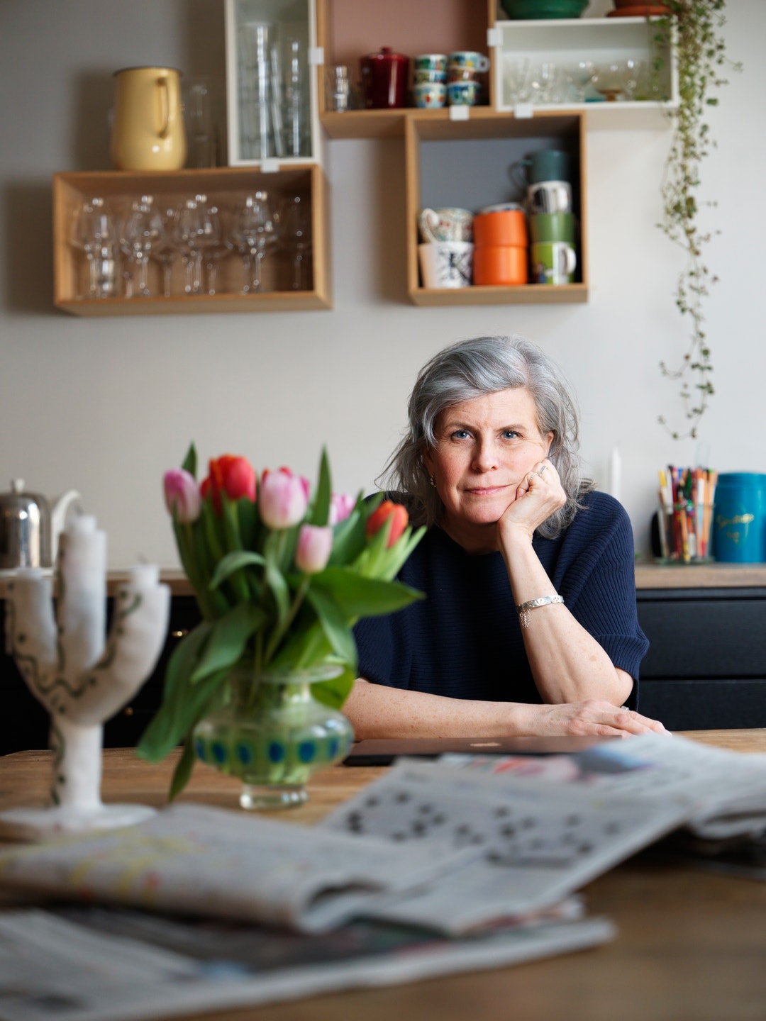 En kvinna sitter vid sitt skrivbord och lutar sitt huvud mot handen. Bakom henne finns väggmonterade hyllor med böcker och dekorationer. På skrivbordet står en vas med röda tulpaner och tidningar.