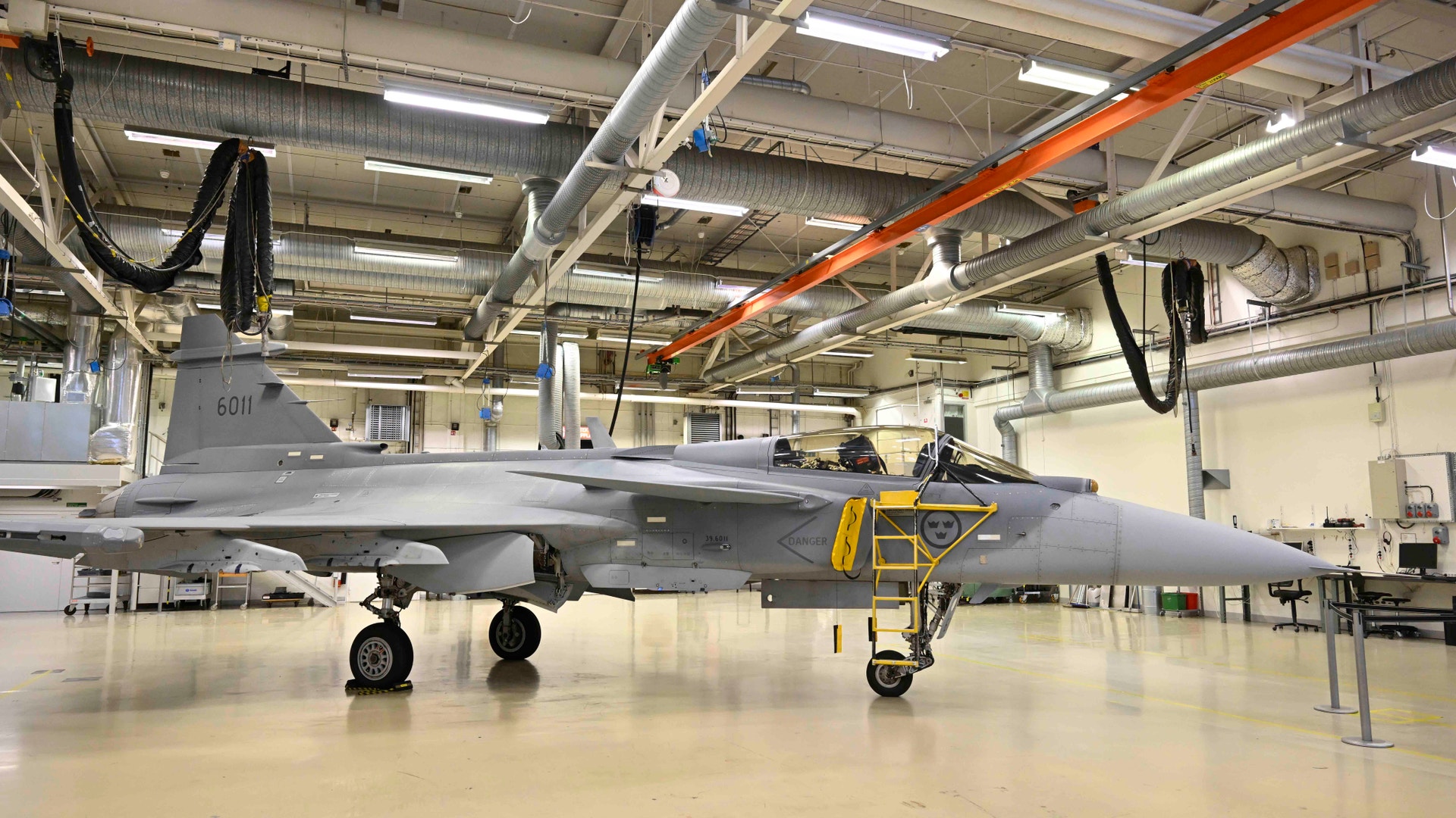 Grått stridsflygplan står parkerat inuti en stor industriell hangar med vita väggar och orange takbalkar.