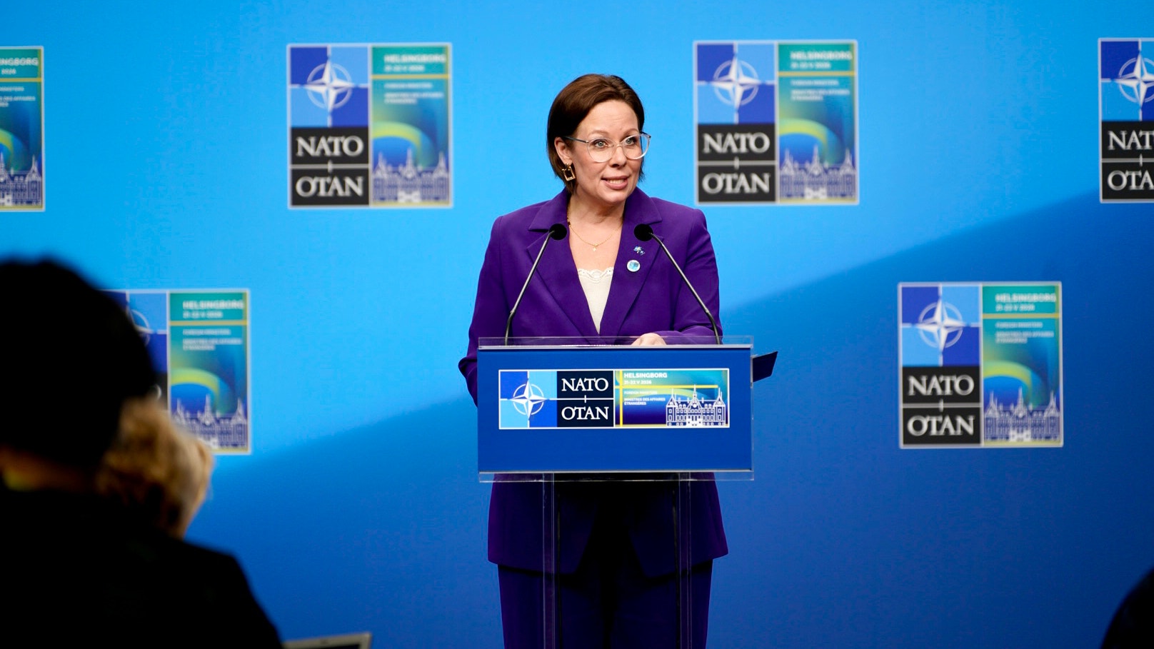 Kvinna i lila kostym talar vid NATO-podium framför blå vägg med NATO-logotyper.