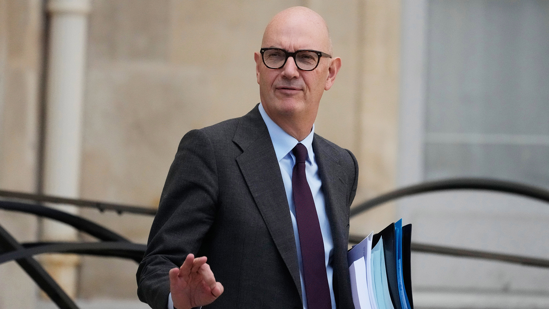 Porträtt av en man i glasögon och mörk kostym med slips, som håller i en mapp och ler mot kameran.