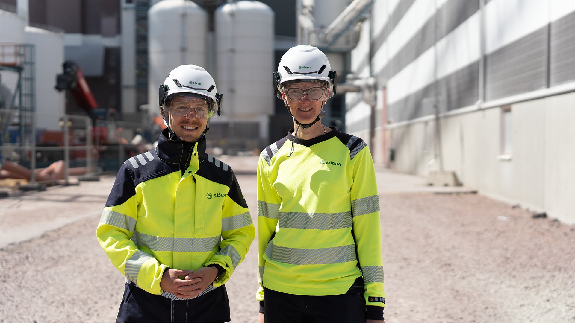 Två arbetare på en byggarbetsplats. De bär vita skyddshjälmar och gula reflekterande västar. I bakgrunden syns industriella byggnader och utrustning.