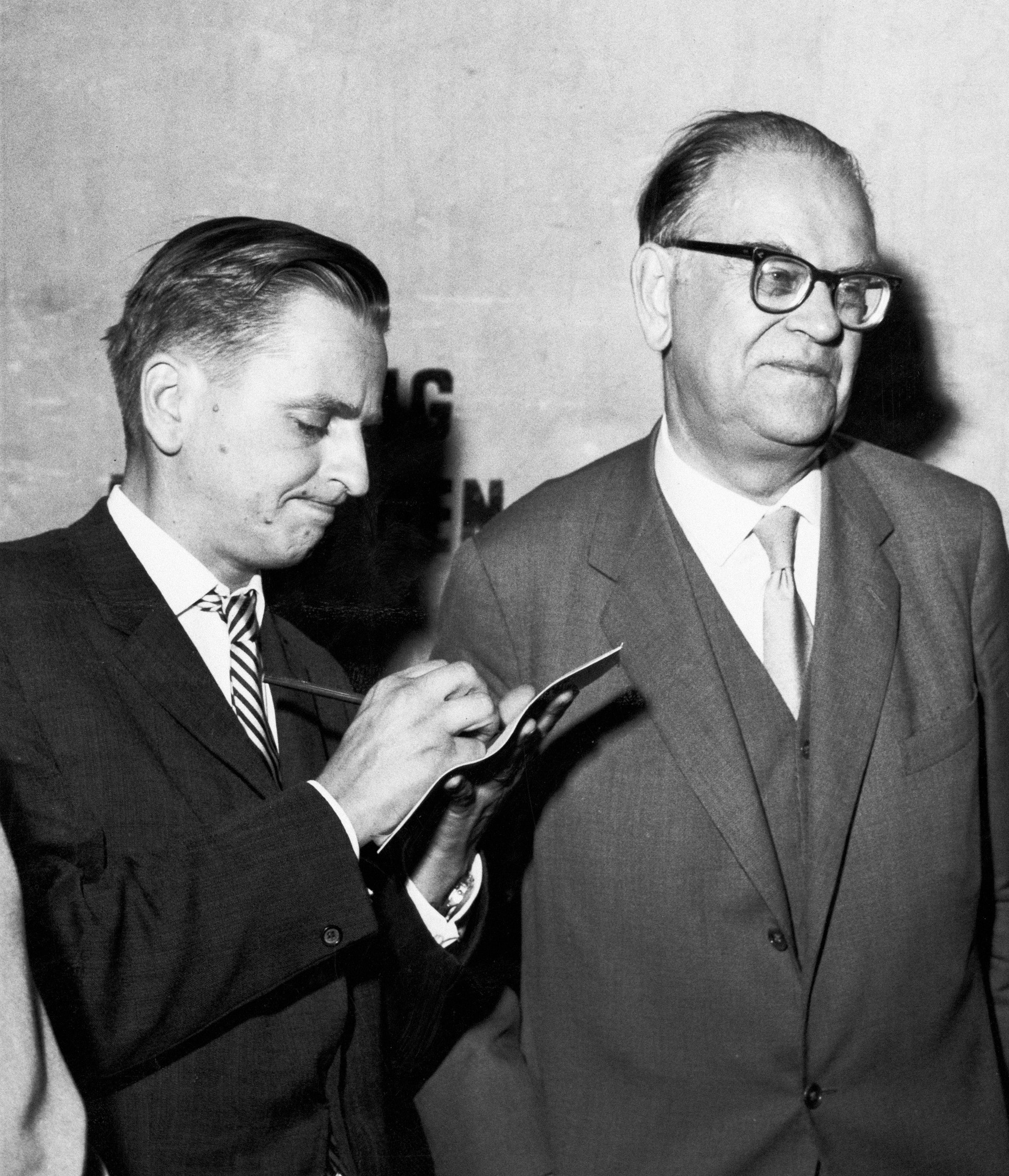Svartvit fotografi från 1960-talet som visar två män i kostym. Mannen till vänster antecknar något på ett block medan mannen till höger med glasögon står bredvid.