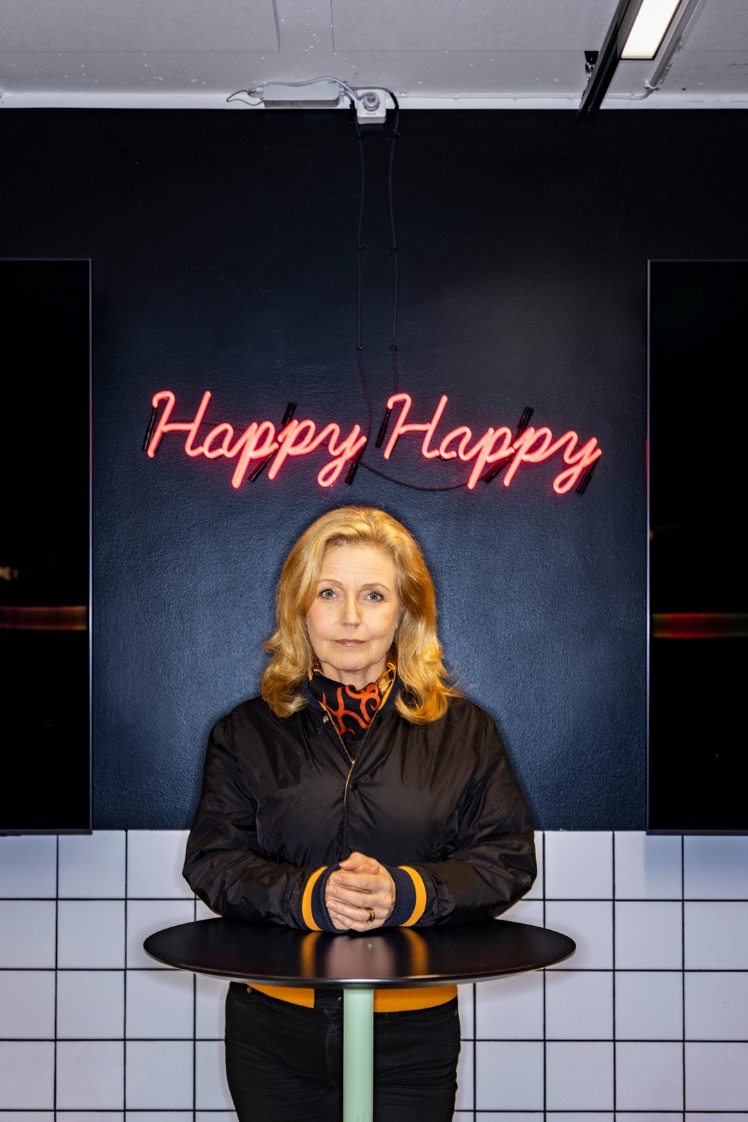 Porträtt av en kvinna framför en mörk vägg med rosa neontext som säger 'Happy Happy'