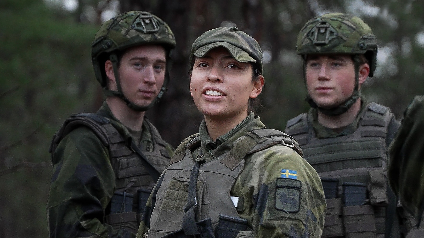 Tre soldater i militär uniform och hjälm. En kvinna i mitten ler, med två män på båda sidor.