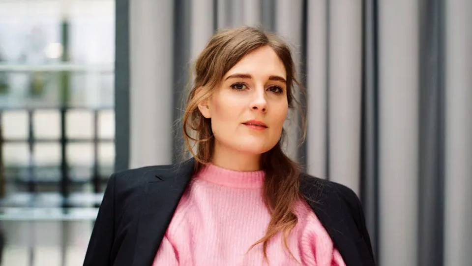 Porträtt av en ung kvinna med långt brunt hår, klädd i en rosa ribbad tröja under en svart blazer. Hon står framför ett fönster med grå gardiner och tittar bort från kameran med ett lugnt, eftertänksamt uttryck.