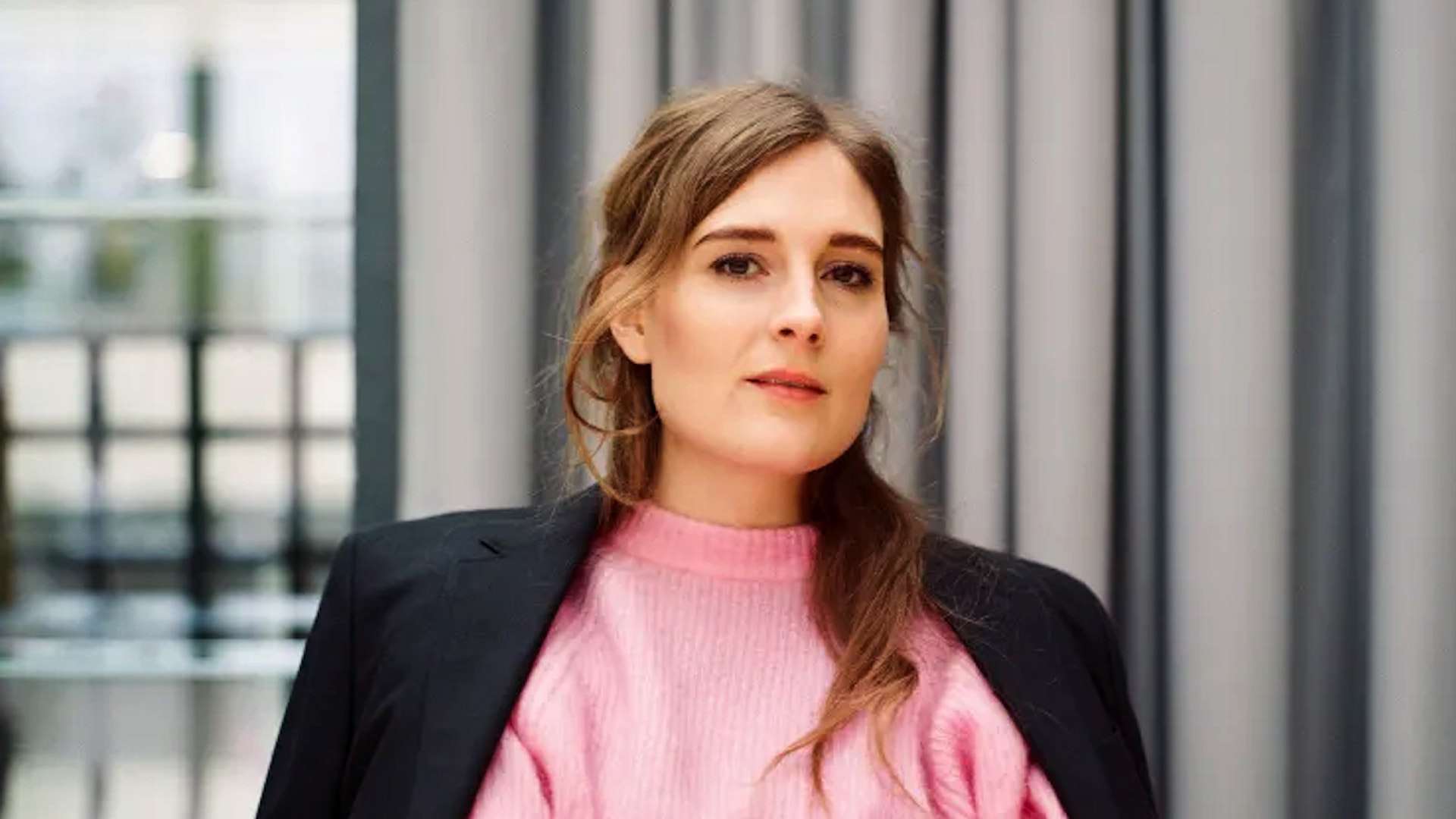 Porträtt av en ung kvinna med långt brunt hår, klädd i en rosa ribbad tröja under en svart blazer. Hon står framför ett fönster med grå gardiner och tittar bort från kameran med ett lugnt, eftertänksamt uttryck.