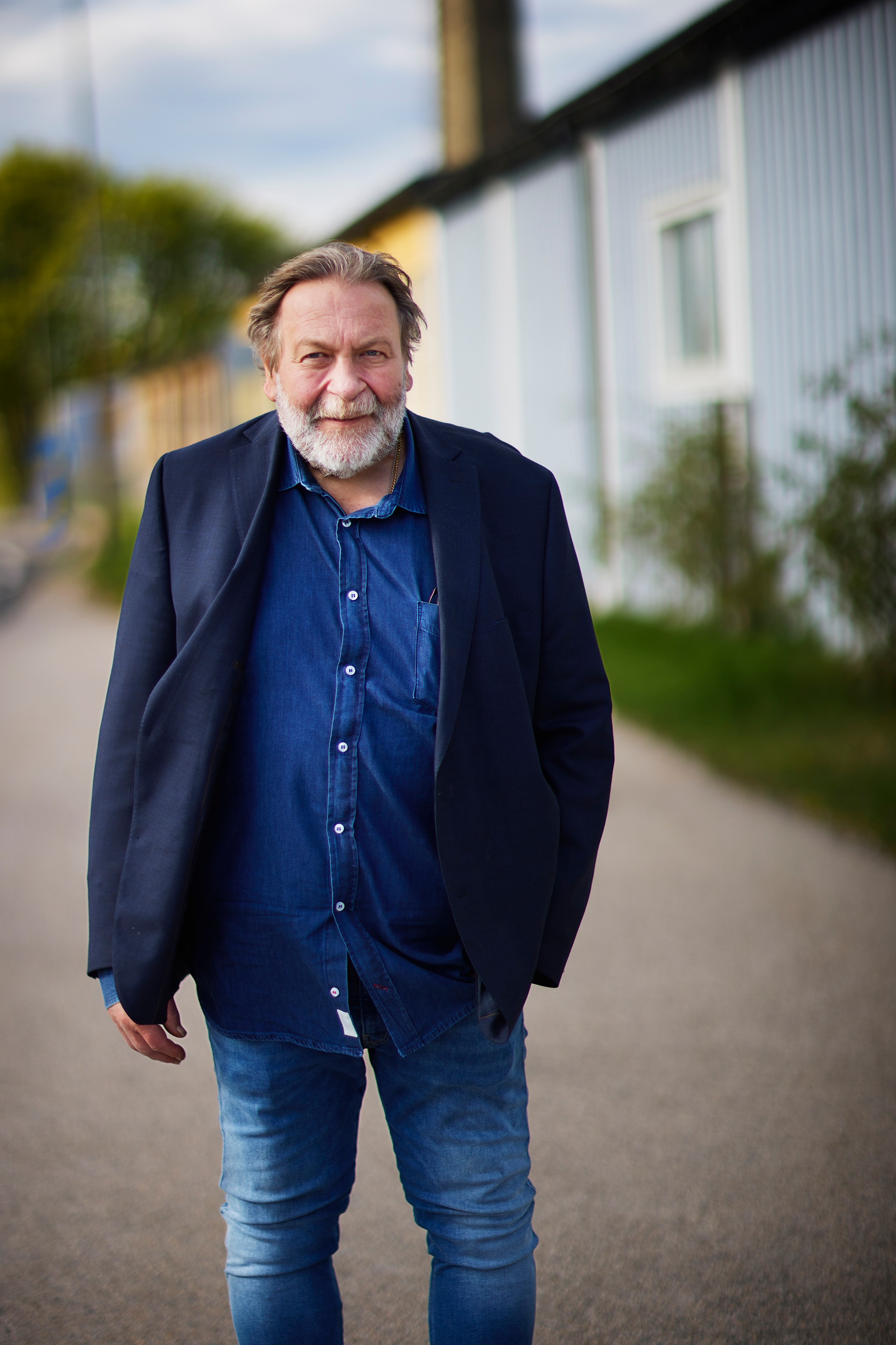 En man med grått hår och skägg står på en väg utomhus. Han bär en mörkblå kavaj över en jeansblå skjorta och jeans. I bakgrunden syns moderna byggnader och gröna växter.