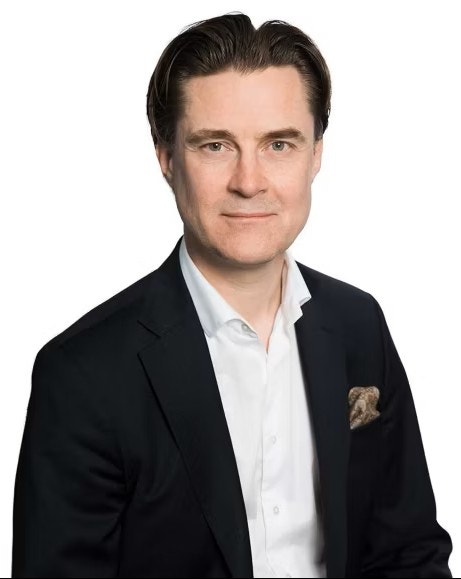 Porträtt av en man i mörk kavaj och vit skjorta mot vit bakgrund.