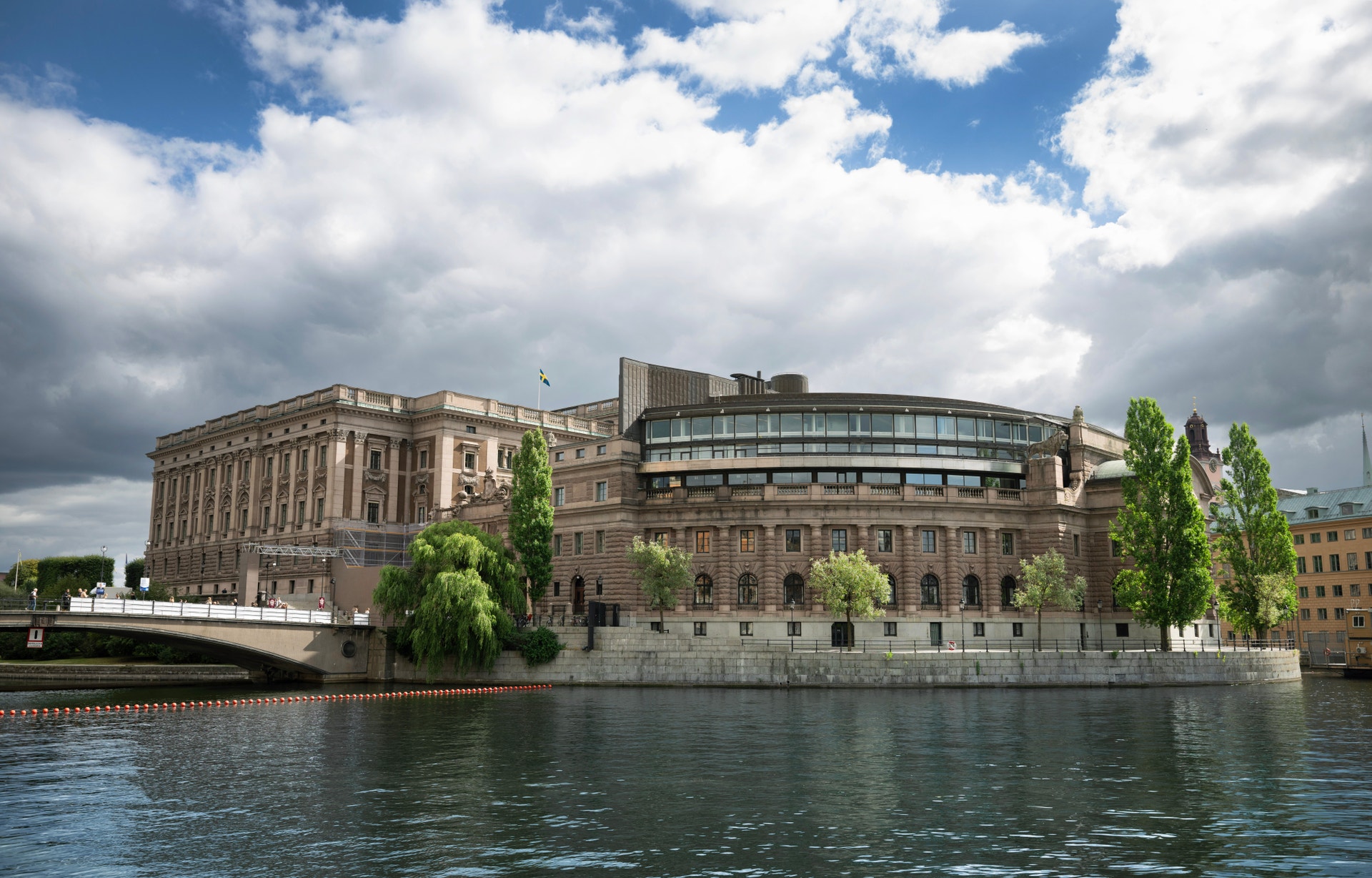 Riksdagshuset i Stockholm ligger vid vattnet. Byggnaden har klassisk gul stenarkitektur med moderna tilbyggnader. Träd växer framför huset och himlen är blå med vita moln.