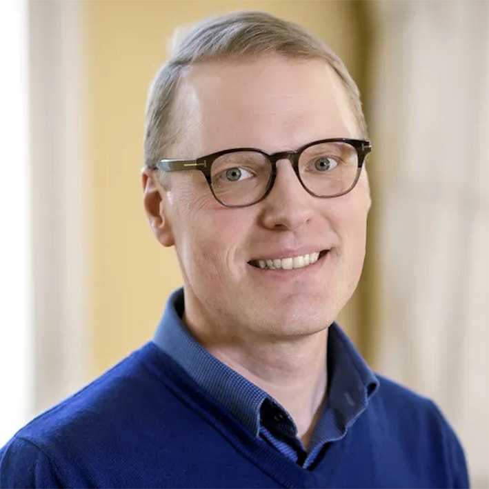 Porträtt av Fredrik NG Andersson, en man med glasögon som ler mot kameran. Han bär en blå tröja och fotografiet är taget mot en gul bakgrund.
