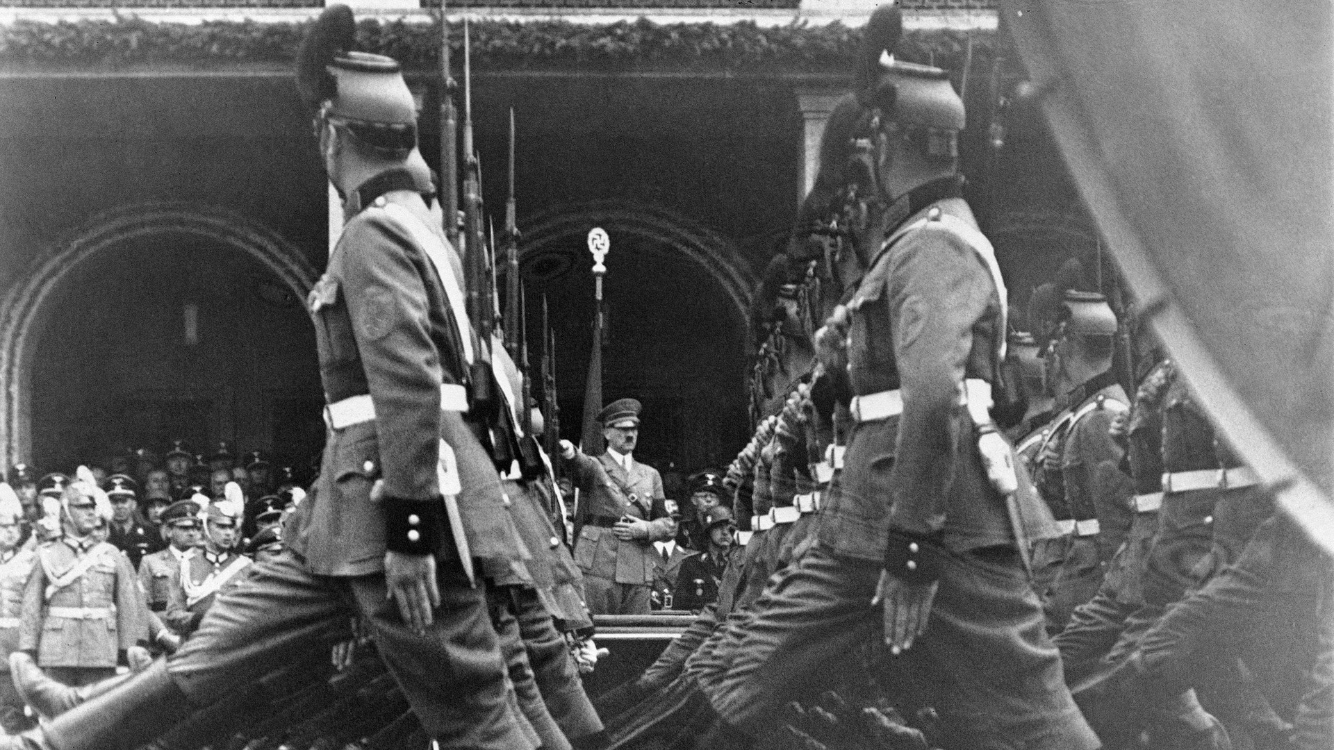 Svartvit historisk bild av soldater som marscherar i paradformation, med åskådare i bakgrunden.