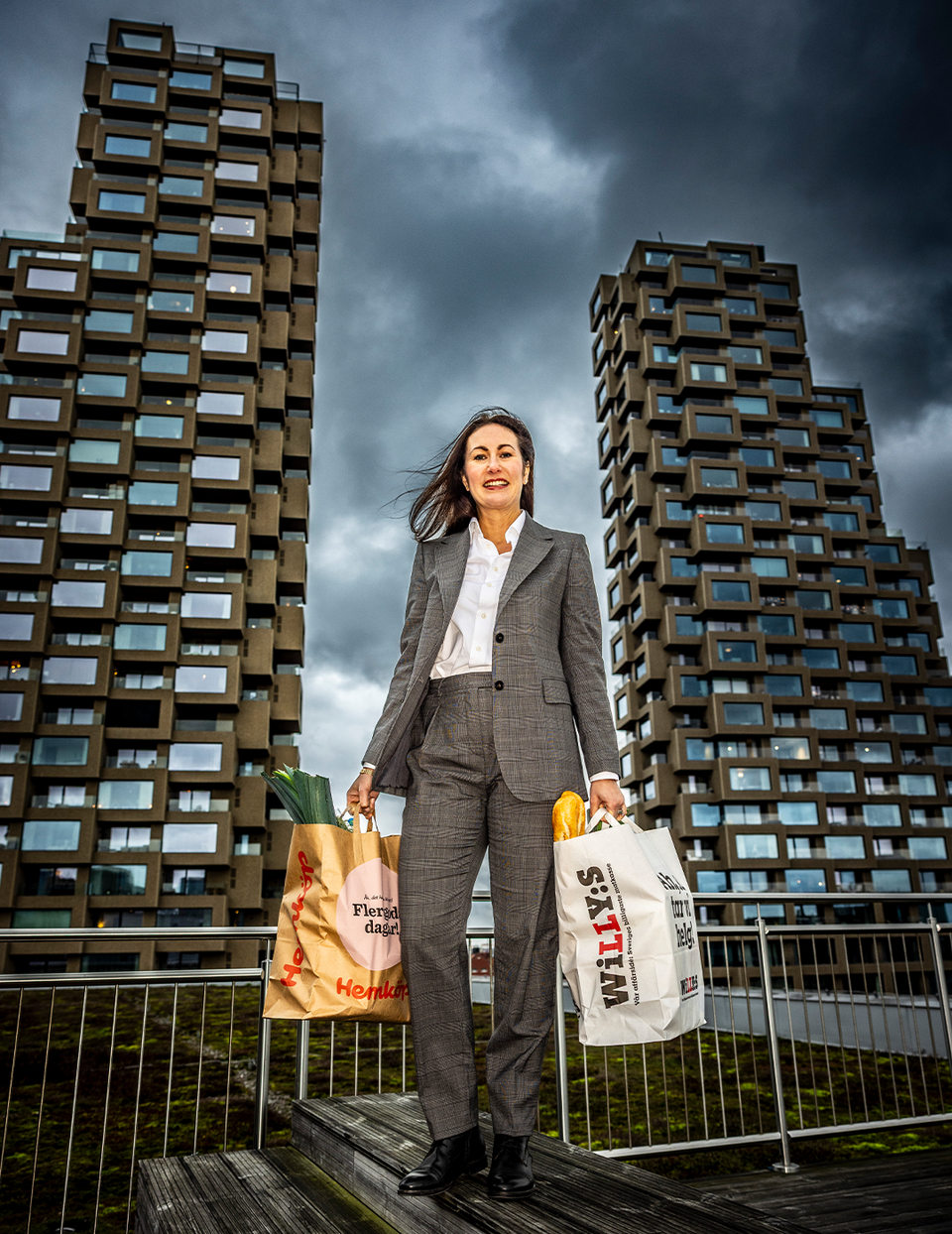 En affärskvinna i grå kostym och vit skjorta står på en trädäck framför två moderna höghus med många fönster. Hon håller två shoppingkassar - en orange och en vit - och ser självsäker ut. Himlen är molnig och dramatisk. En metallräcke syns i förgrunden.