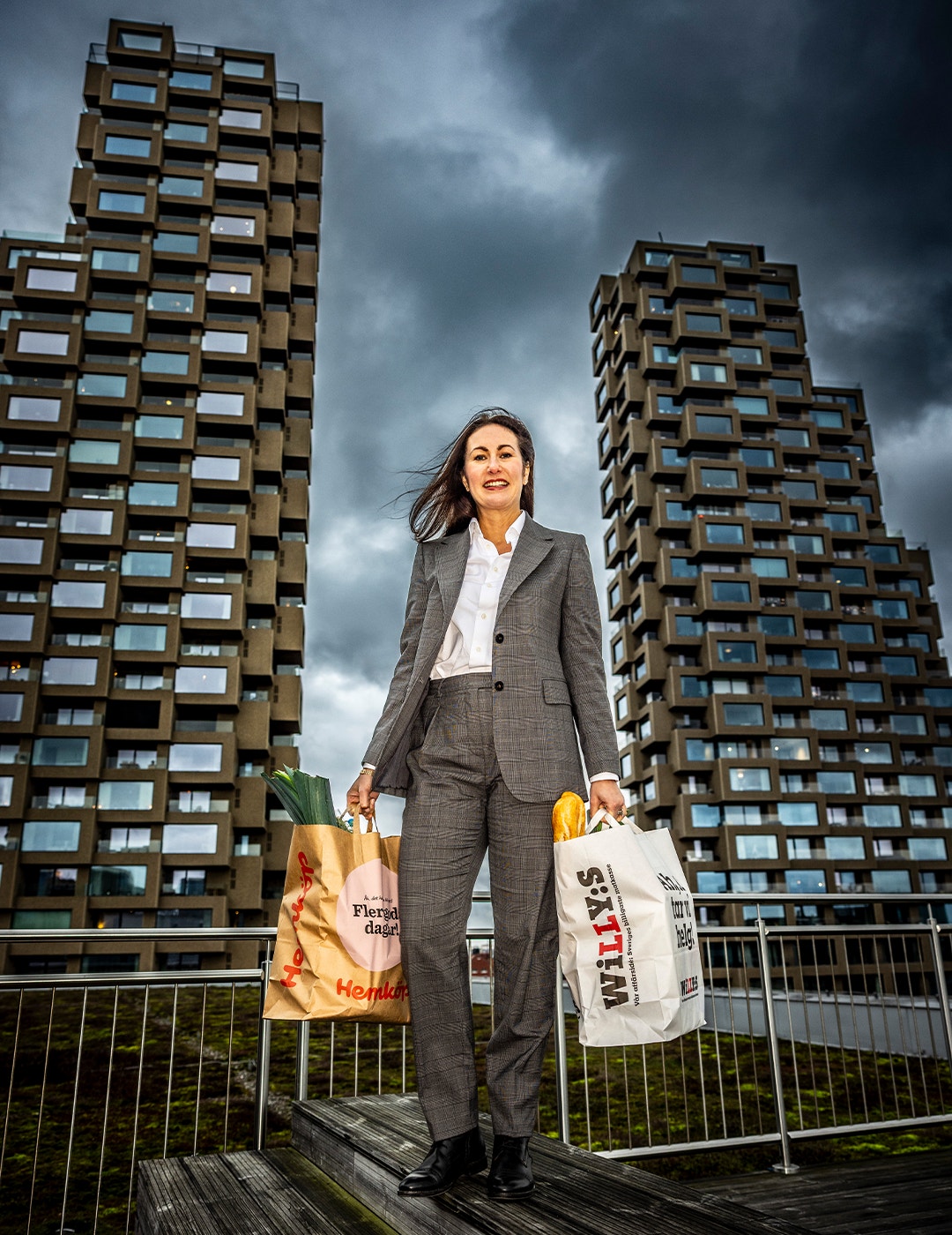 En affärskvinna i grå kostym och vit skjorta står på en trädäck framför två moderna höghus med många fönster. Hon håller två shoppingkassar - en orange och en vit - och ser självsäker ut. Himlen är molnig och dramatisk. En metallräcke syns i förgrunden.