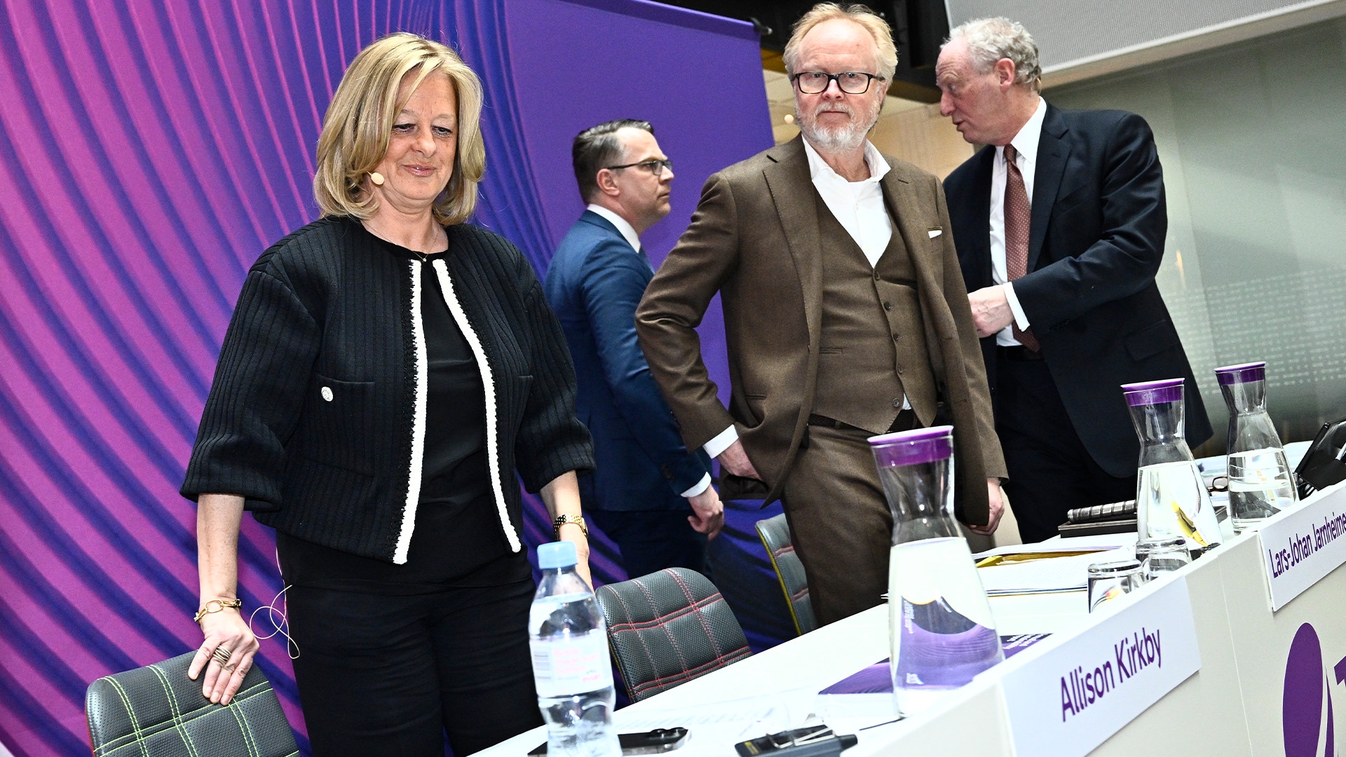 Fem personer i affärskläder står vid ett bord under en konferens eller paneldiskussion. En lila bakgrund med vertikala streck syns bakom dem.