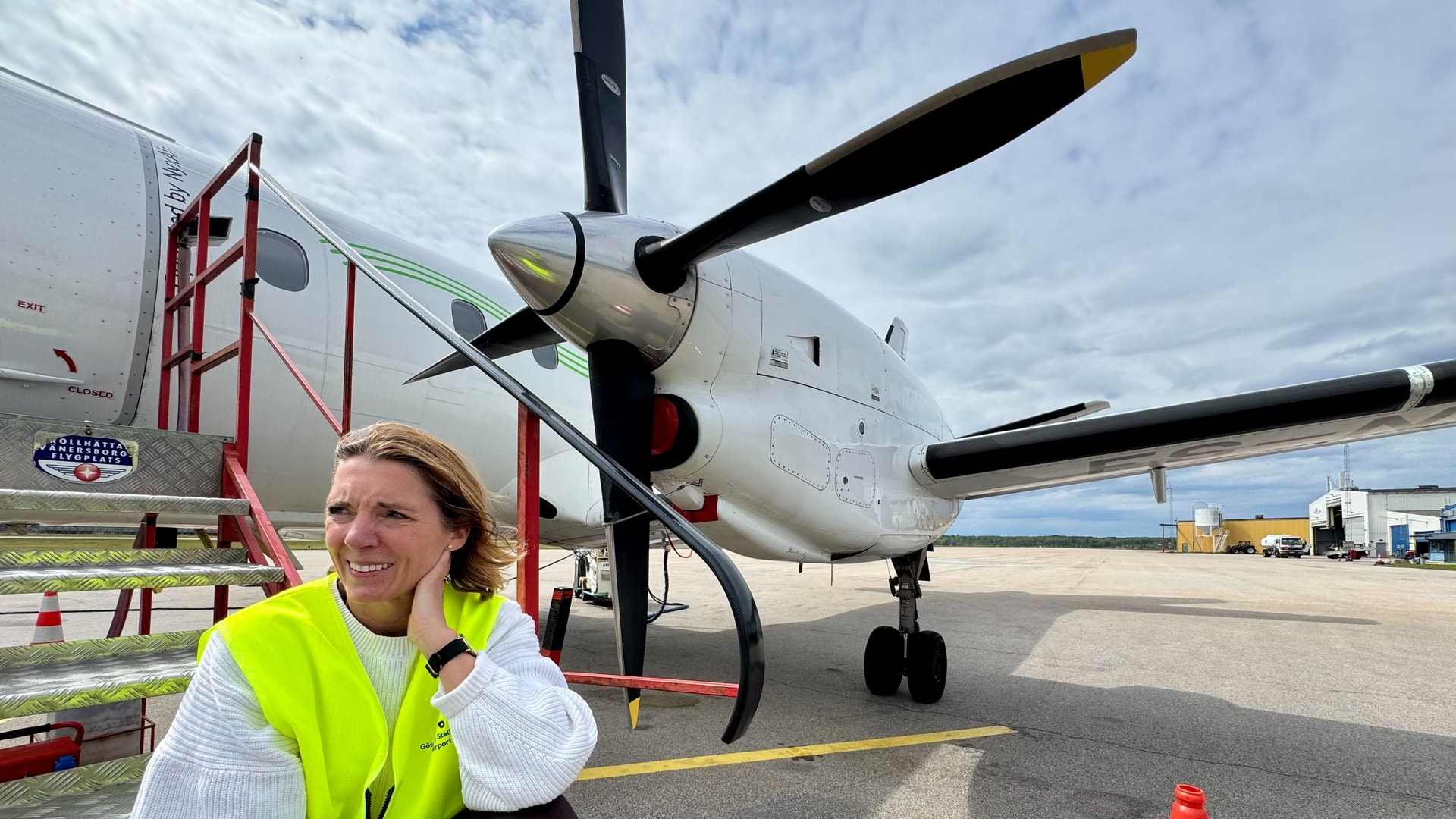 Kvinna i gul väst sitter framför ett propellerflygplan på en flygplats.