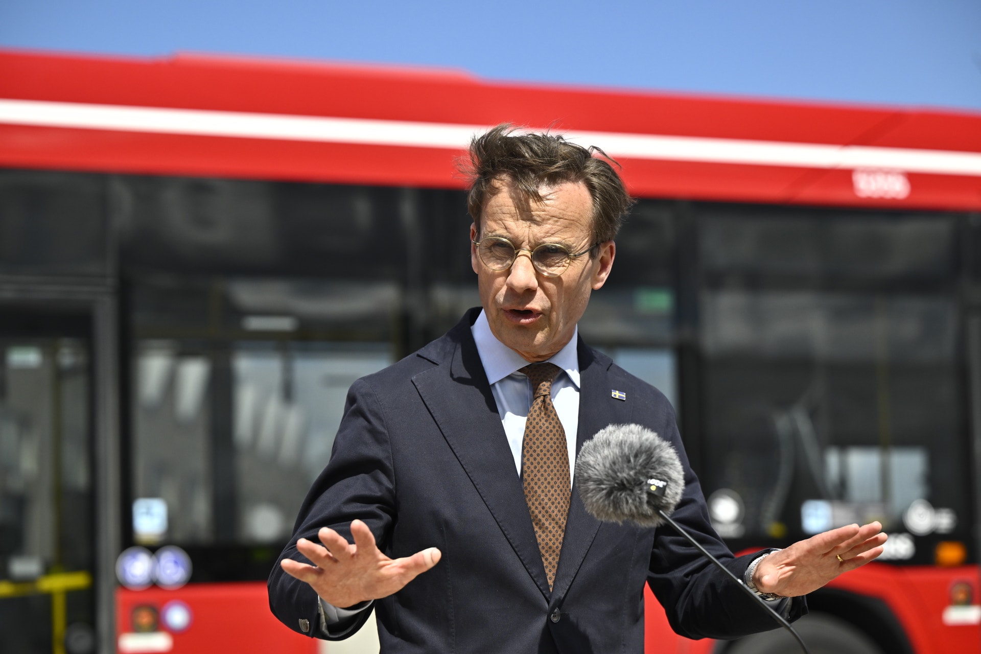 En man i mörk kostym talar vid en mikrofon framför en röd och vit buss.