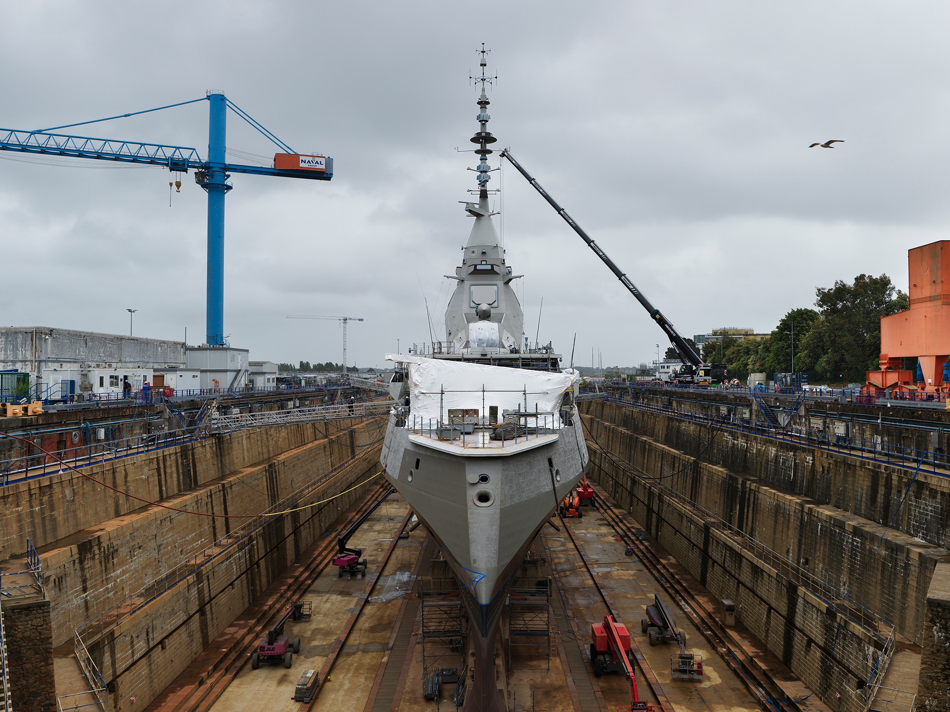 Ett militärt marinfartyg med vit skrov ligger i en skeppsdocka omgiven av höga betongväggar. En stor blå kran syns i bakgrunden tillsammans med andra industriella byggnader.