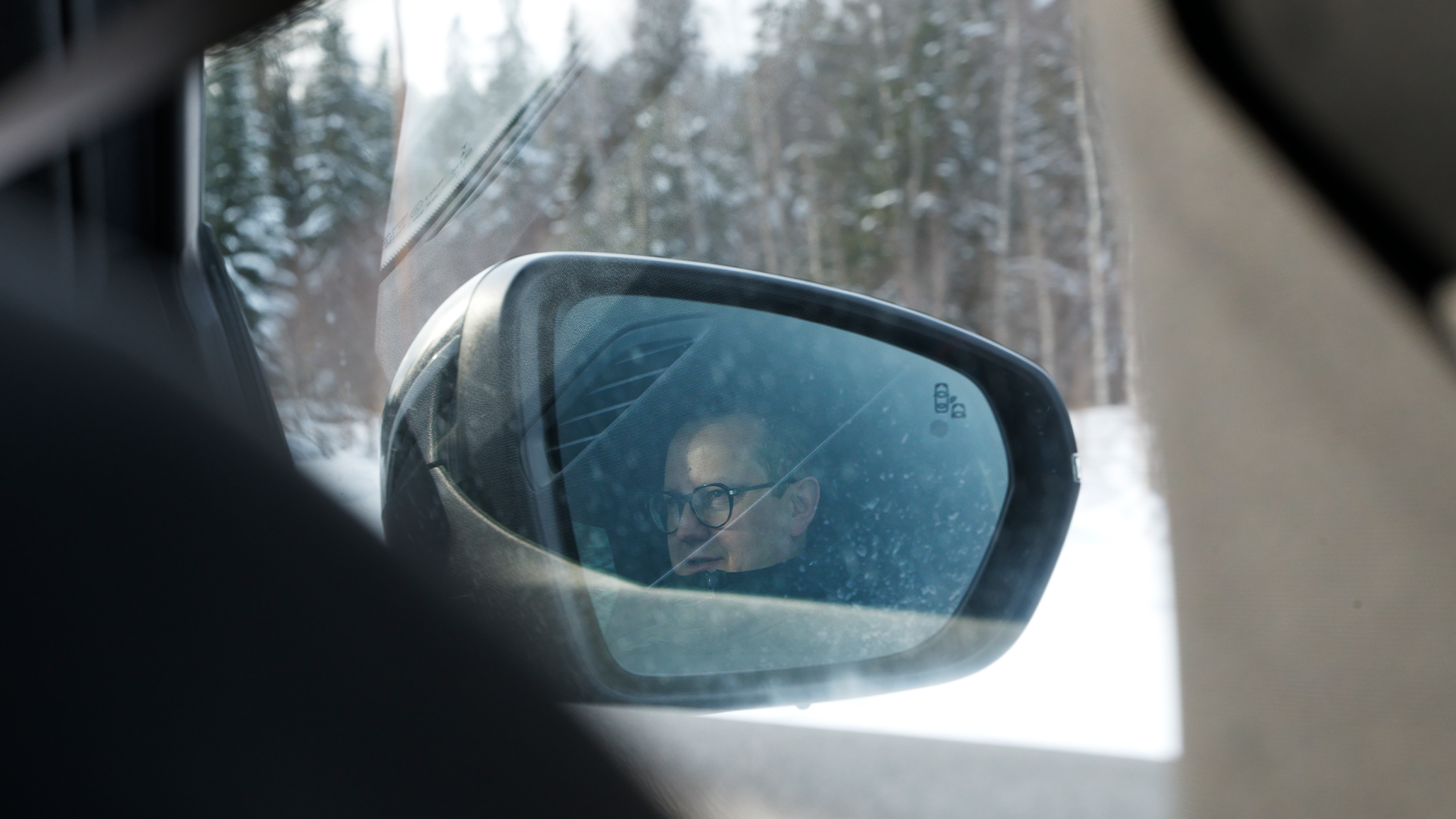 En person reflekterad i en bilspegel med snöig skog i bakgrunden.