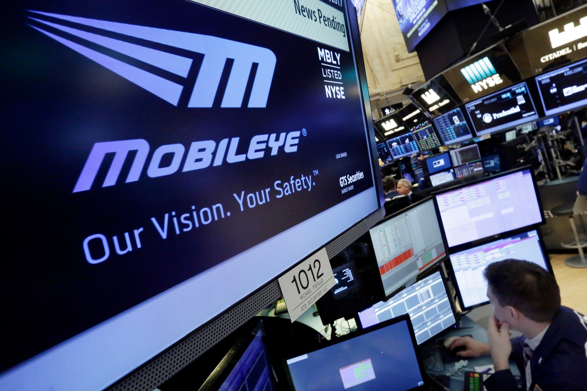 En stor skärm på börsen visar Mobileye-logotypen med texten 'Our Vision. Your Safety.' I bakgrunden arbetar börsmäklare vid handelsterminaler.