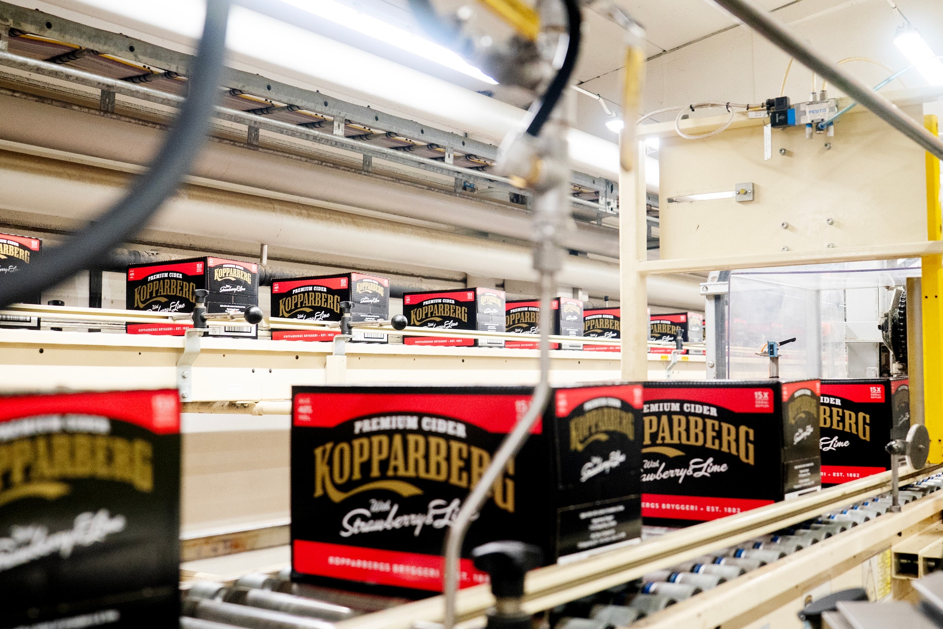 Flaskor med Kopparberg-cider på en produktionslinje i en fabrik. Flaskorna har röda och svarta etiketter och transporteras på ett band. Fabriksutrustning och gula säkerhetselement syns i bakgrunden.
