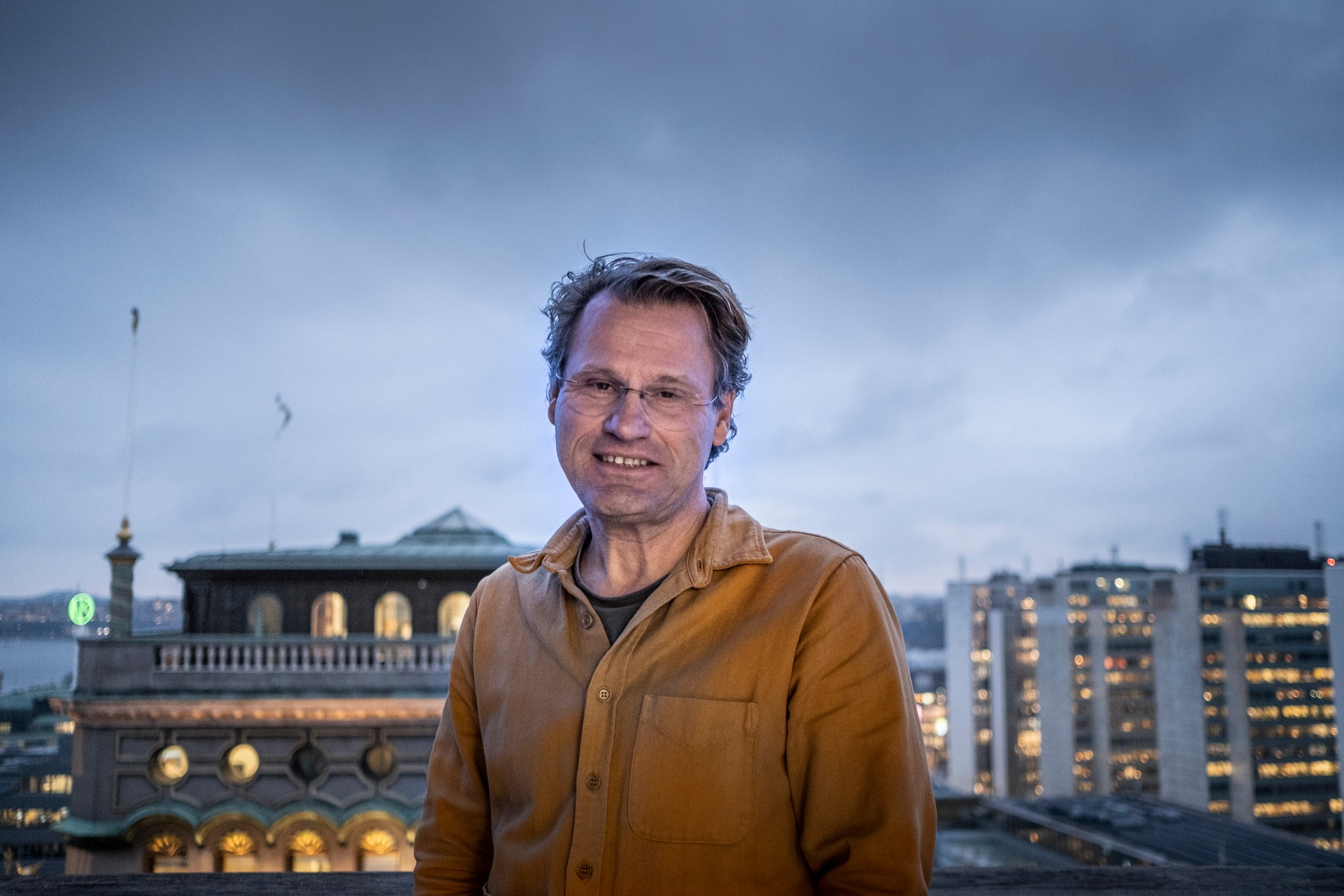Porträtt av en man med glasögon i gul-brun skjorta mot en stadsskyline med belyst byggnader under skymningen.