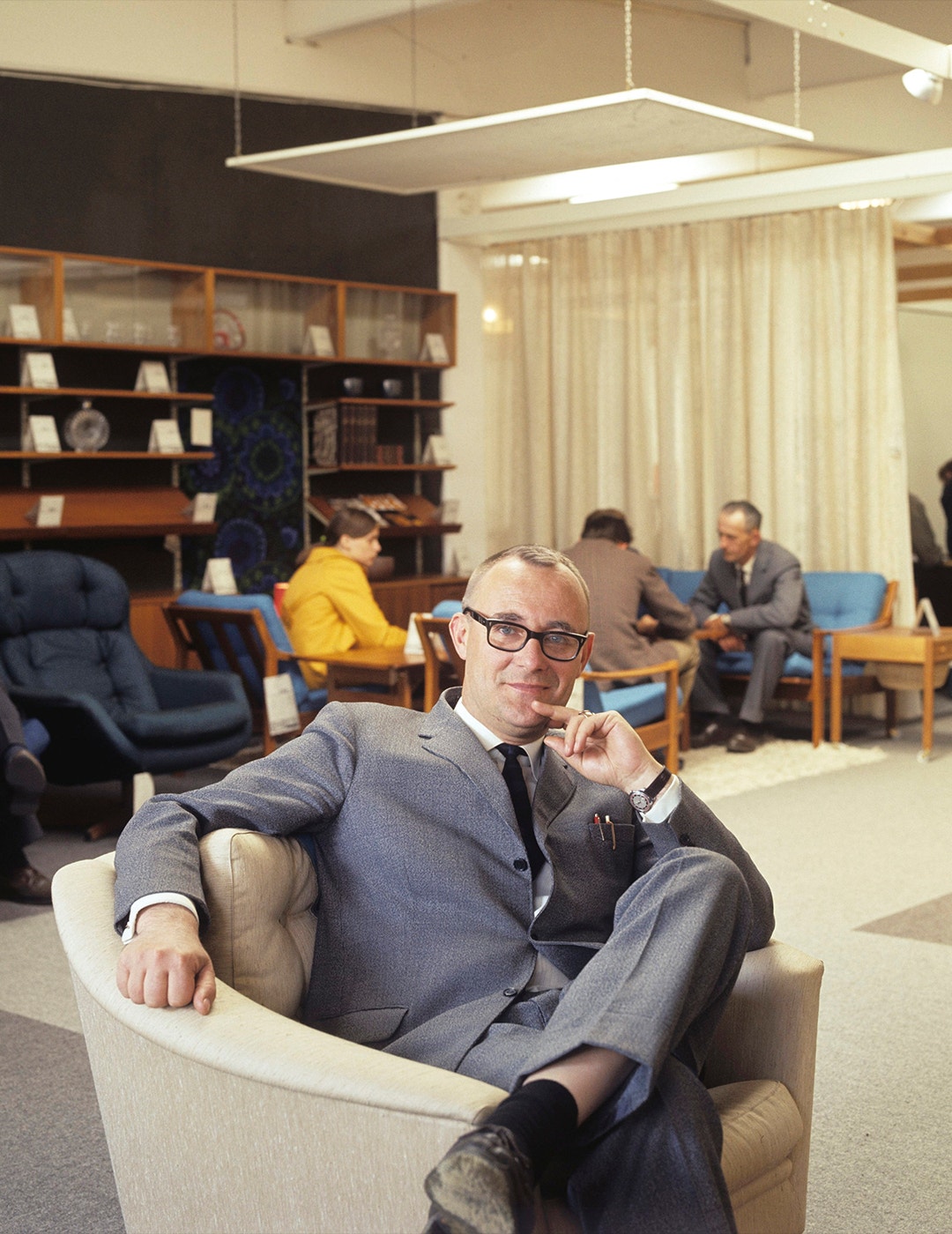 Man i glasögon och kostym sitter i en modern kontorsmiljö med möbler och andra människor i bakgrunden.