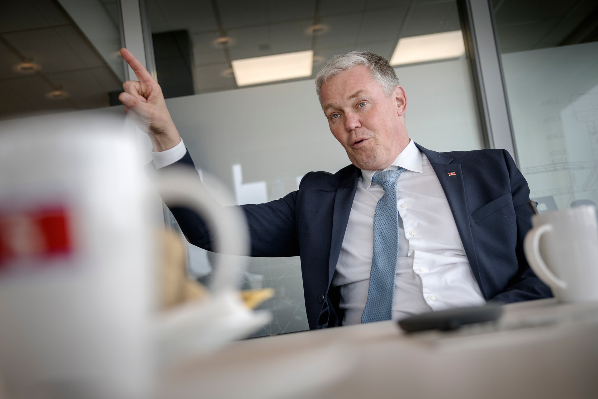 En man i kostym och slips sitter vid ett skrivbord och pratar med handen upplyft, som för att illustrera en poäng.