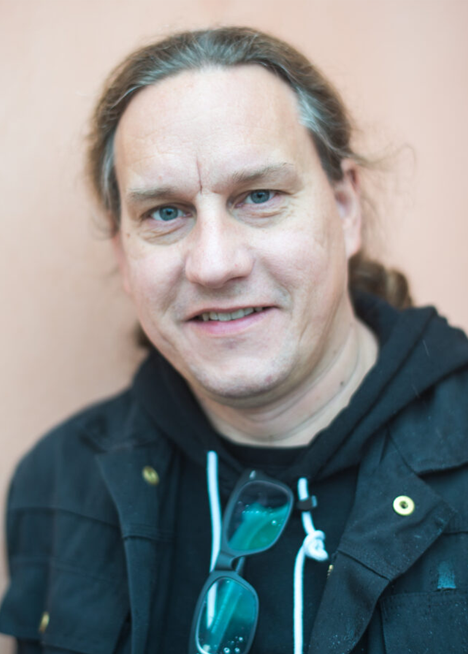 Tommy Andersson tittar in och ler i kameran. Han har på sig en svart hoodie, en svart jacka och ett par glasögon som hänger i kragen. 