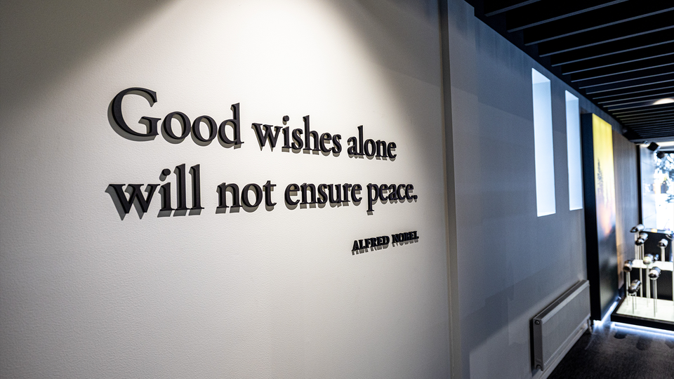 Väggtext med citat "Good wishes alone will not ensure peace" av Alfred Nobel på en vit vägg i ett modernt interiör.