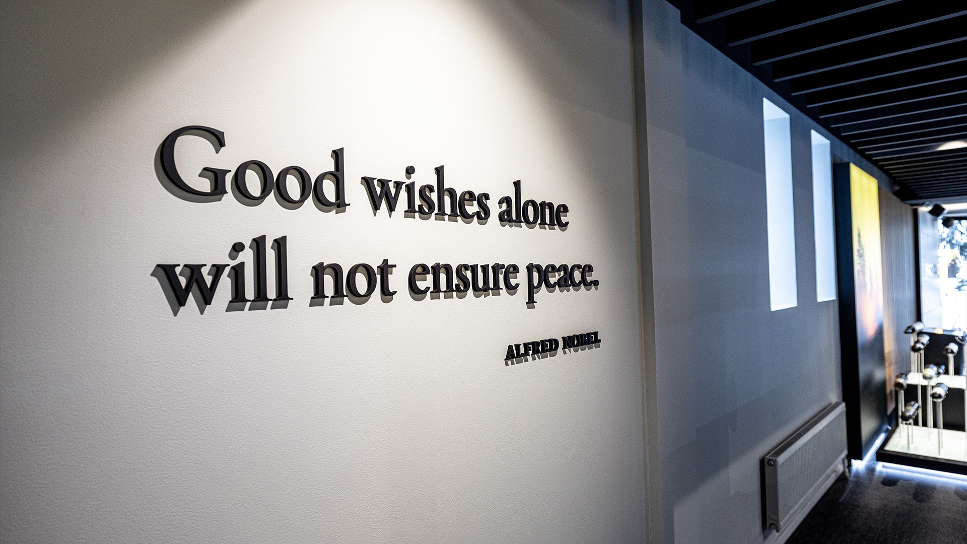 Väggtext med citat "Good wishes alone will not ensure peace" av Alfred Nobel på en vit vägg i ett modernt interiör.