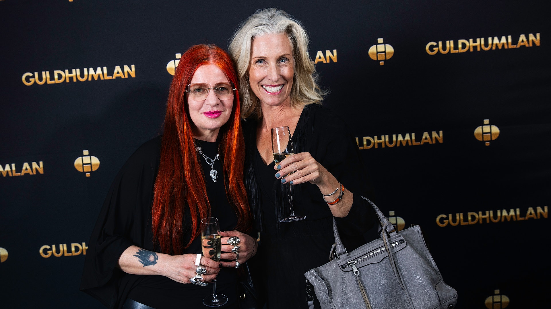 Två kvinnor poserar tillsammans framför en Guldhumlan-bakgrund. De håller champagneglas och ler mot kameran.