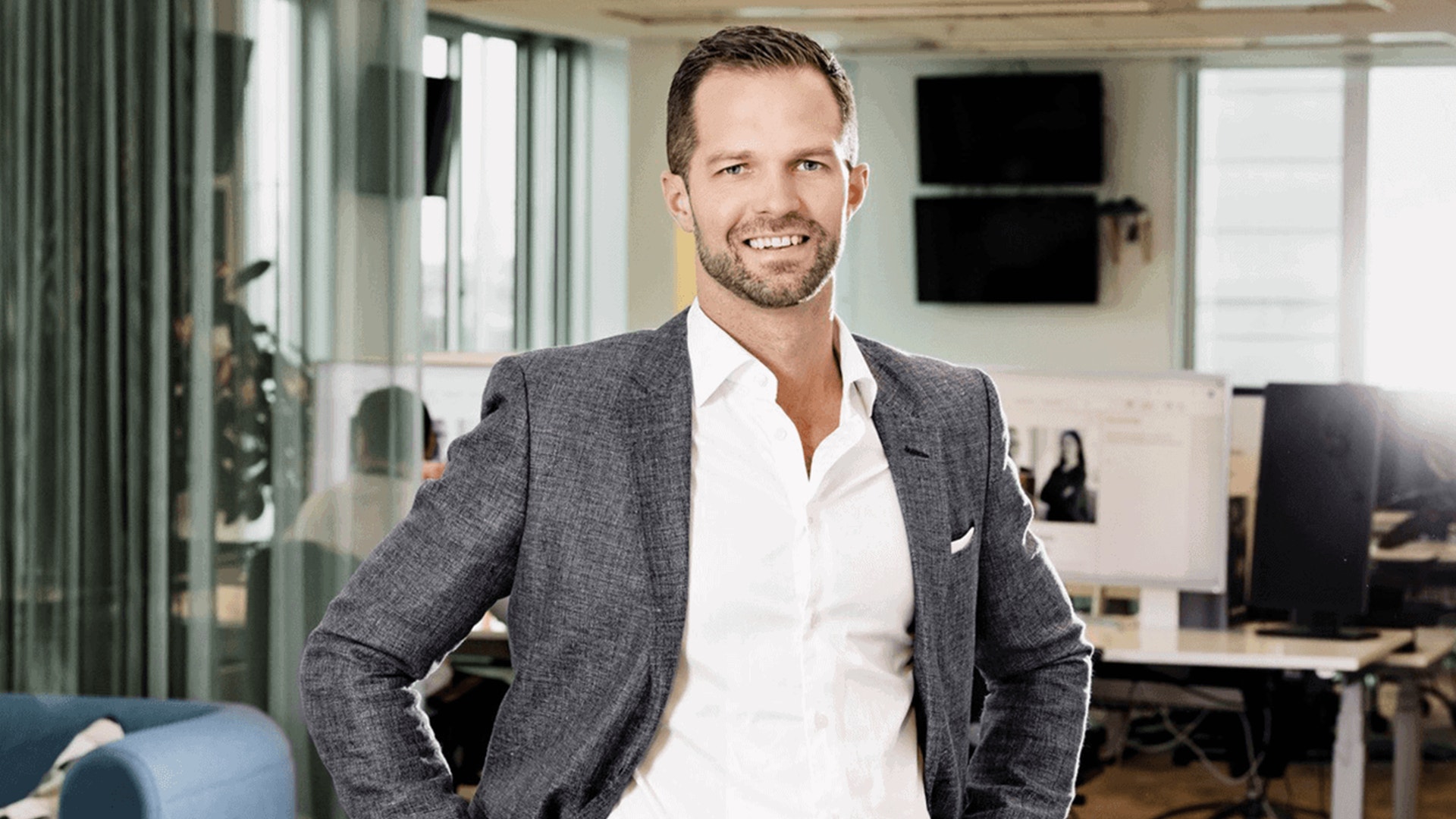 En man i grå blazer och vit skjorta står leende i ett modernt kontor.