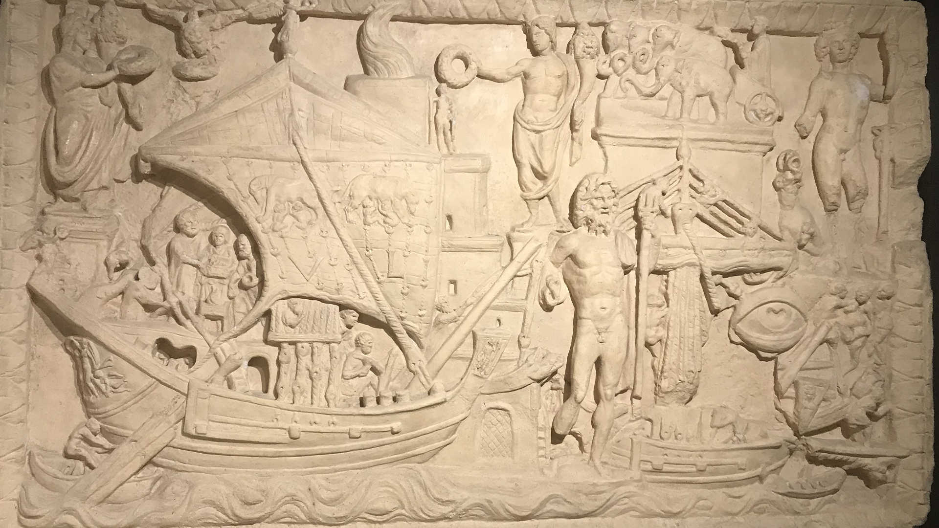 Stenskulptur med relief som visar antika scener med människor, båtar och klassiska motiv från forntiden.