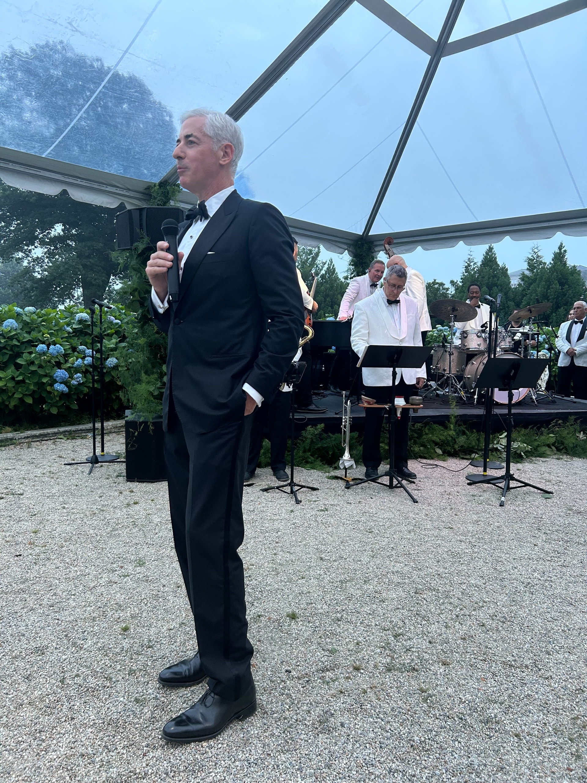Bill Ackman håller tal till gästerna efter tennisturneringen Finance Cup. Han är klädd i en smoking och har ett jazzband bakom sig. 
