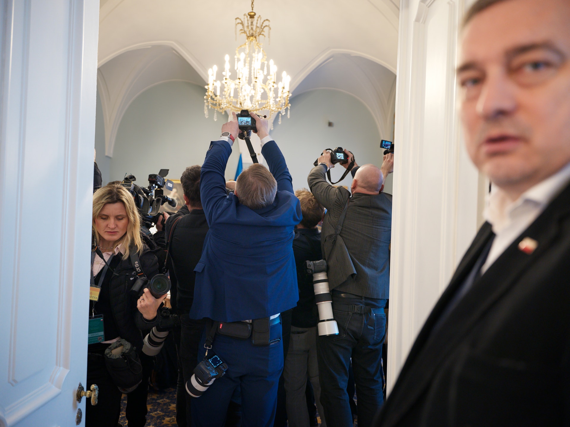 Grupp av människor i en elegant sal med kristallkrona. Flera personer fotograferar under vad som verkar vara ett officiellt evenemang.