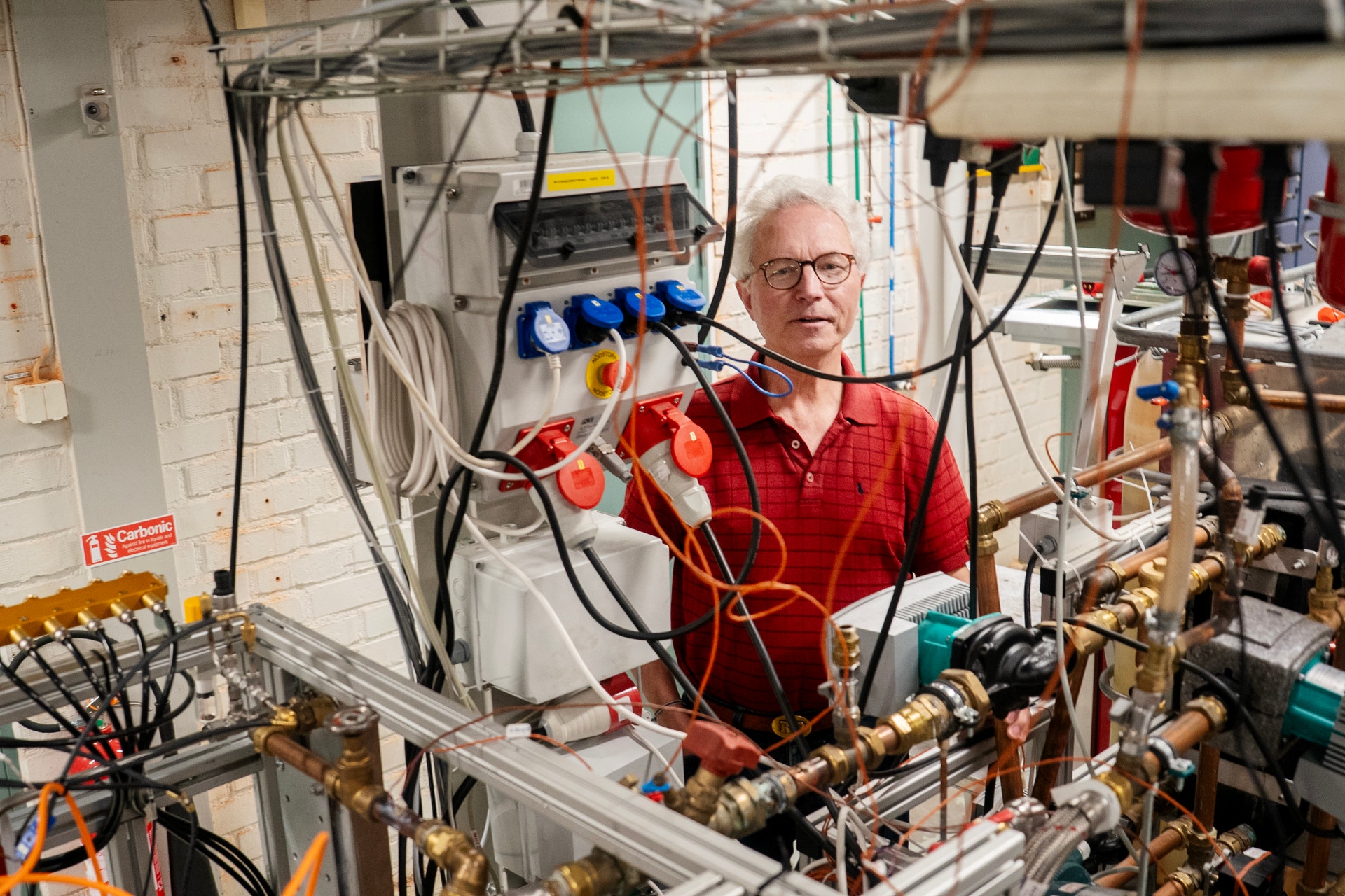 En man i röd skjorta och glasögon står i ett laboratorium omgiven av elektronisk utrustning och kablar.