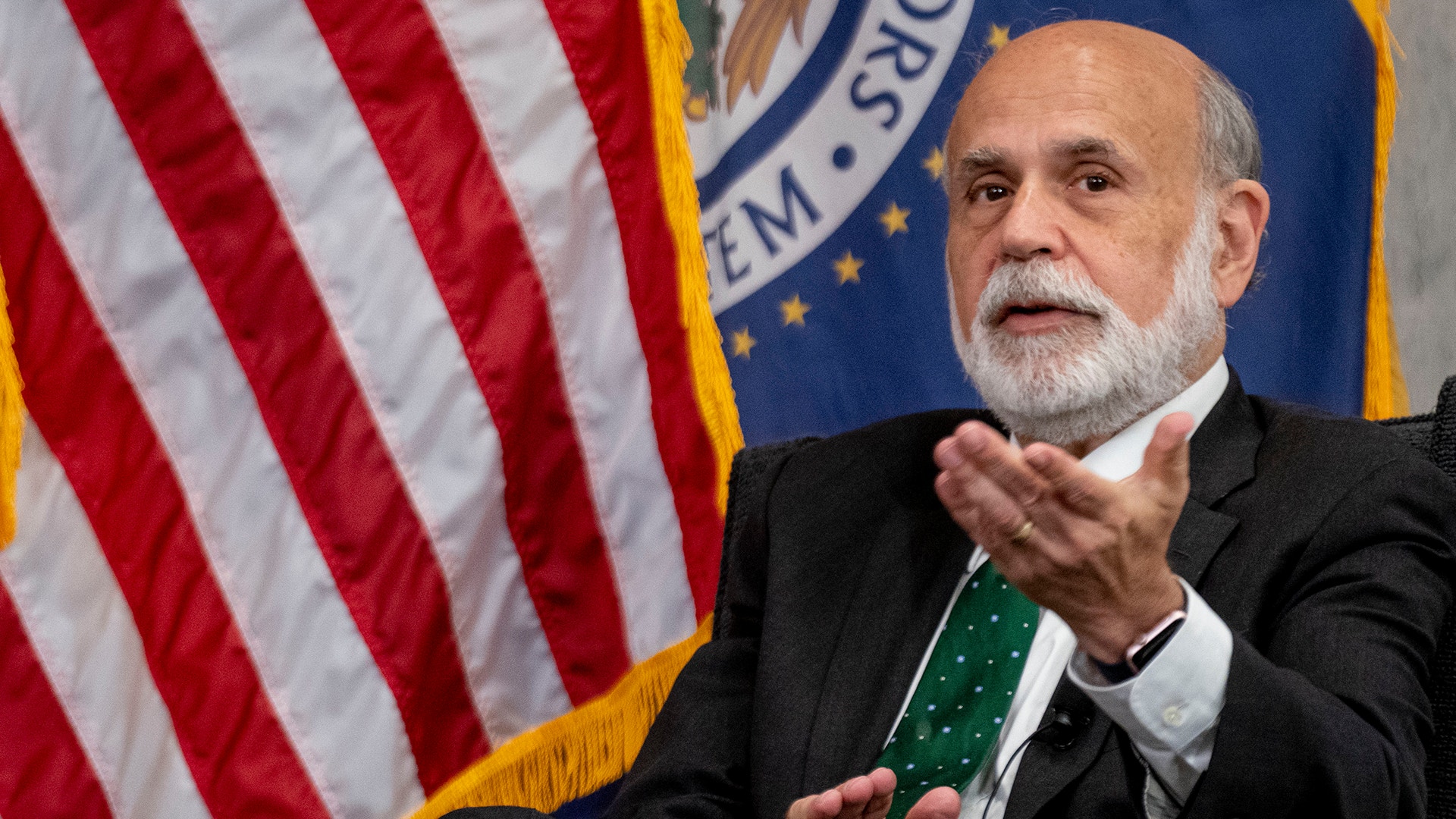 Porträtt av en man i kostym som talar vid ett officiellt evenemang med amerikansk flagga i bakgrunden.