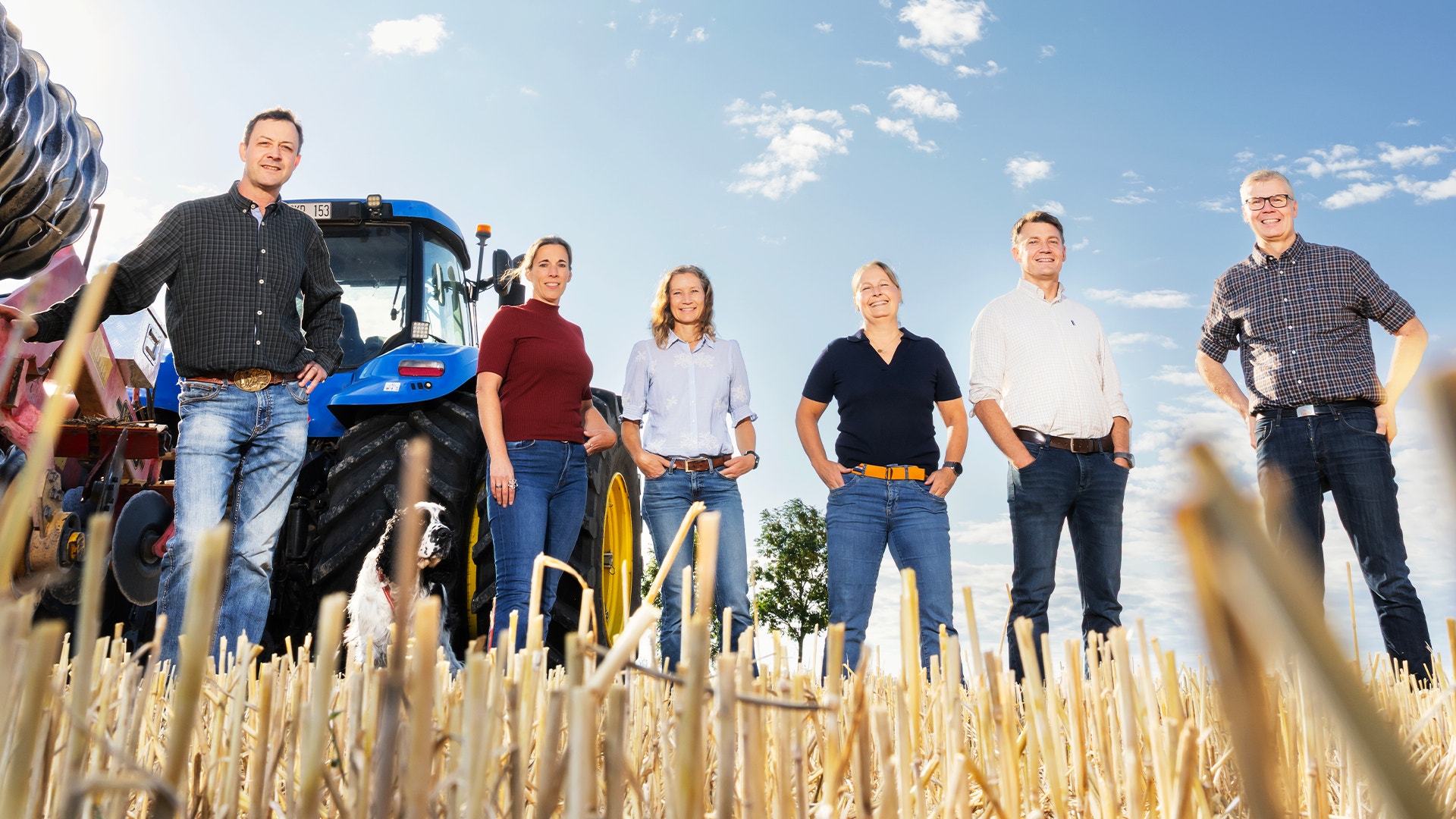 Sex vuxna människor står på ett stubbåker framför en blå traktor en solig dag.