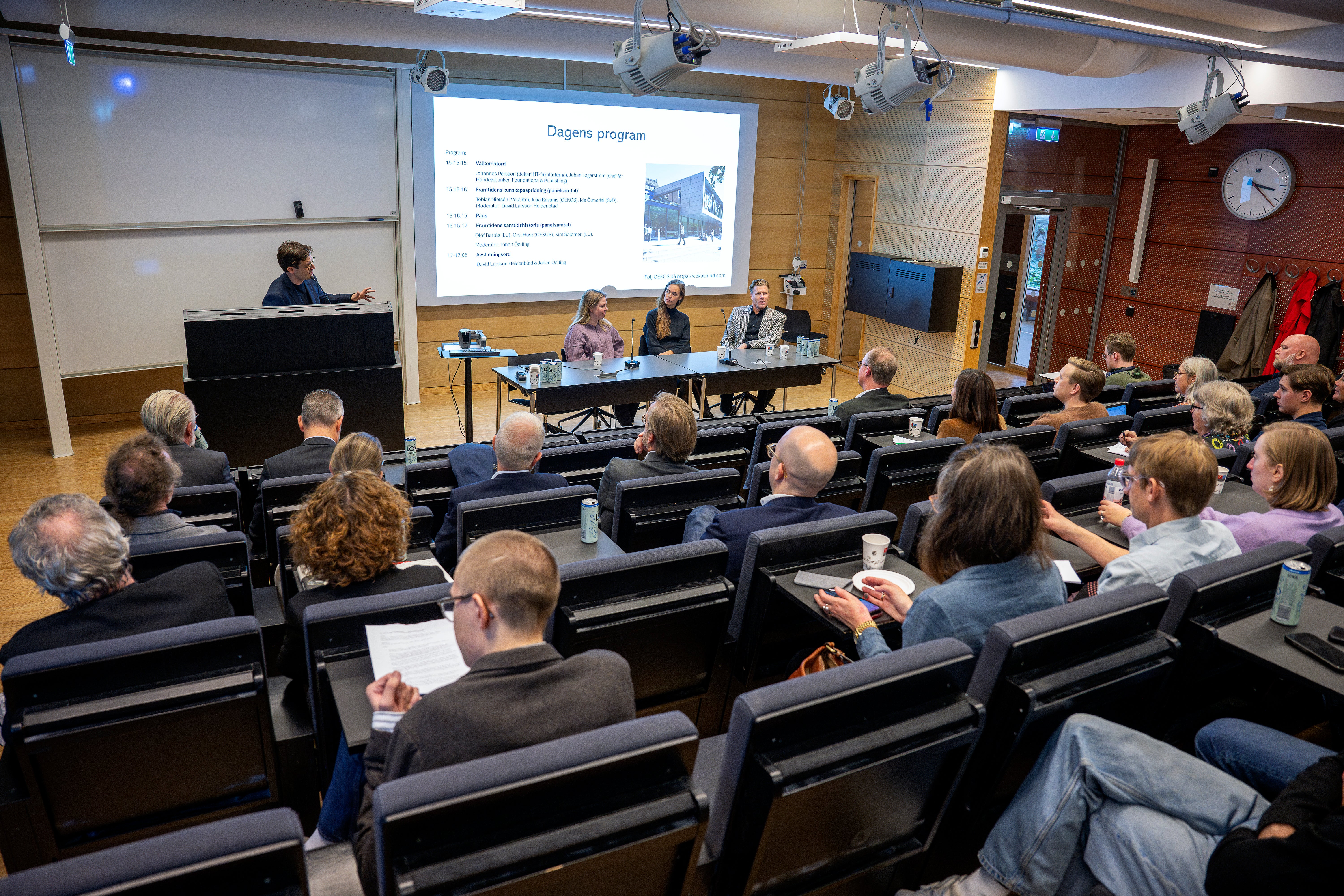 Publik sitter i en föreläsningssal och lyssnar på ett panelsamtal. Längst fram sitter tre personer vid ett bord, och en person talar vid ett podium bredvid en projektorduk med rubriken Dagens program.
