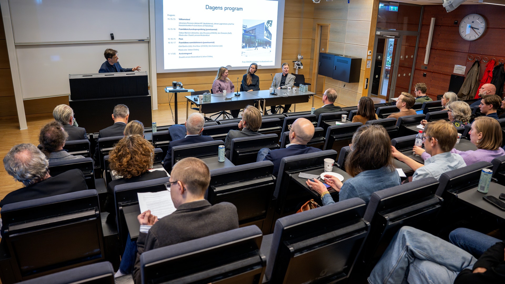Publik sitter i en föreläsningssal och lyssnar på ett panelsamtal. Längst fram sitter tre personer vid ett bord, och en person talar vid ett podium bredvid en projektorduk med rubriken Dagens program.