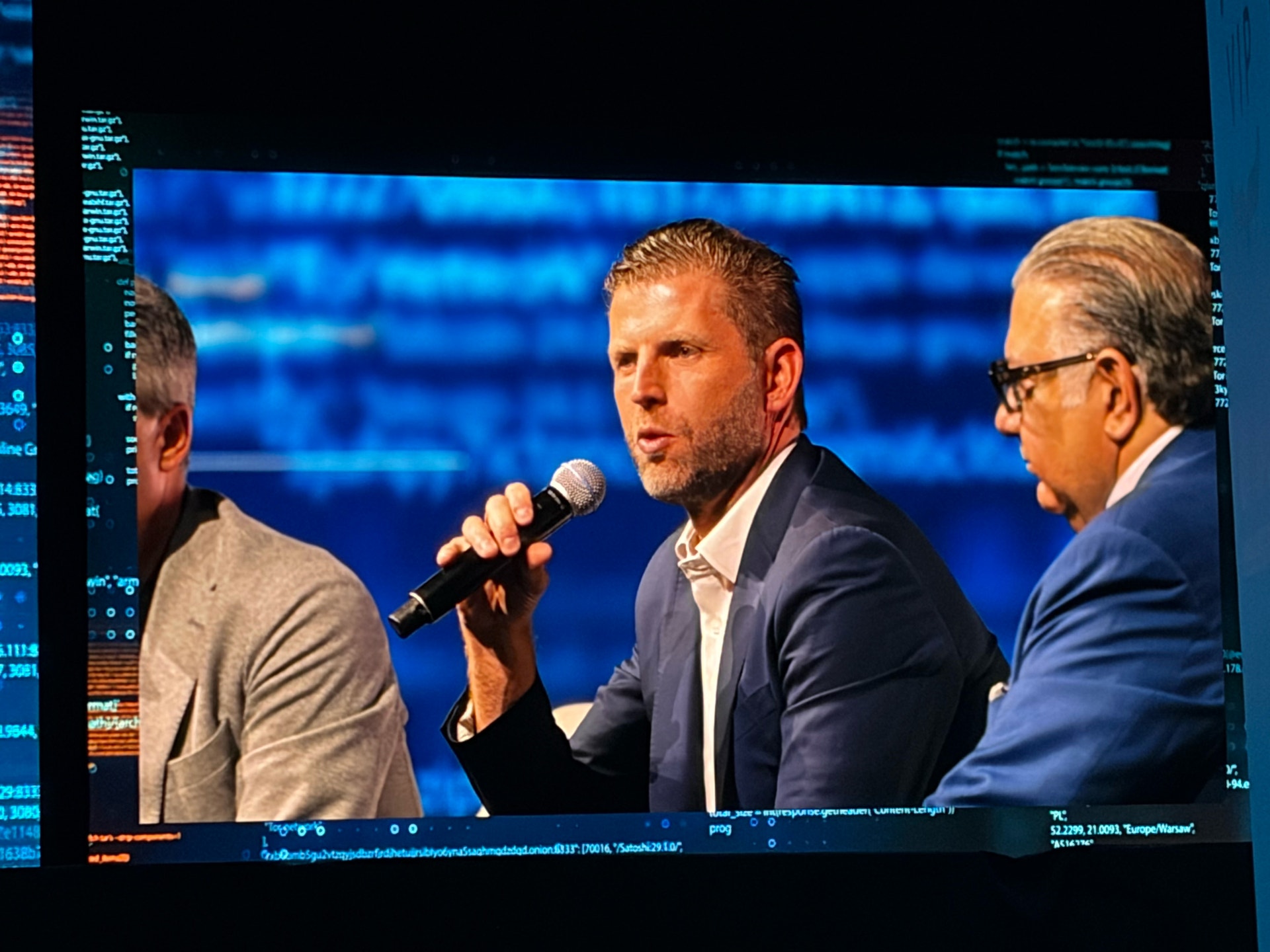 Tre män på en scen under en konferens. Mannen i mitten talar i en mikrofon medan två andra män lyssnar.