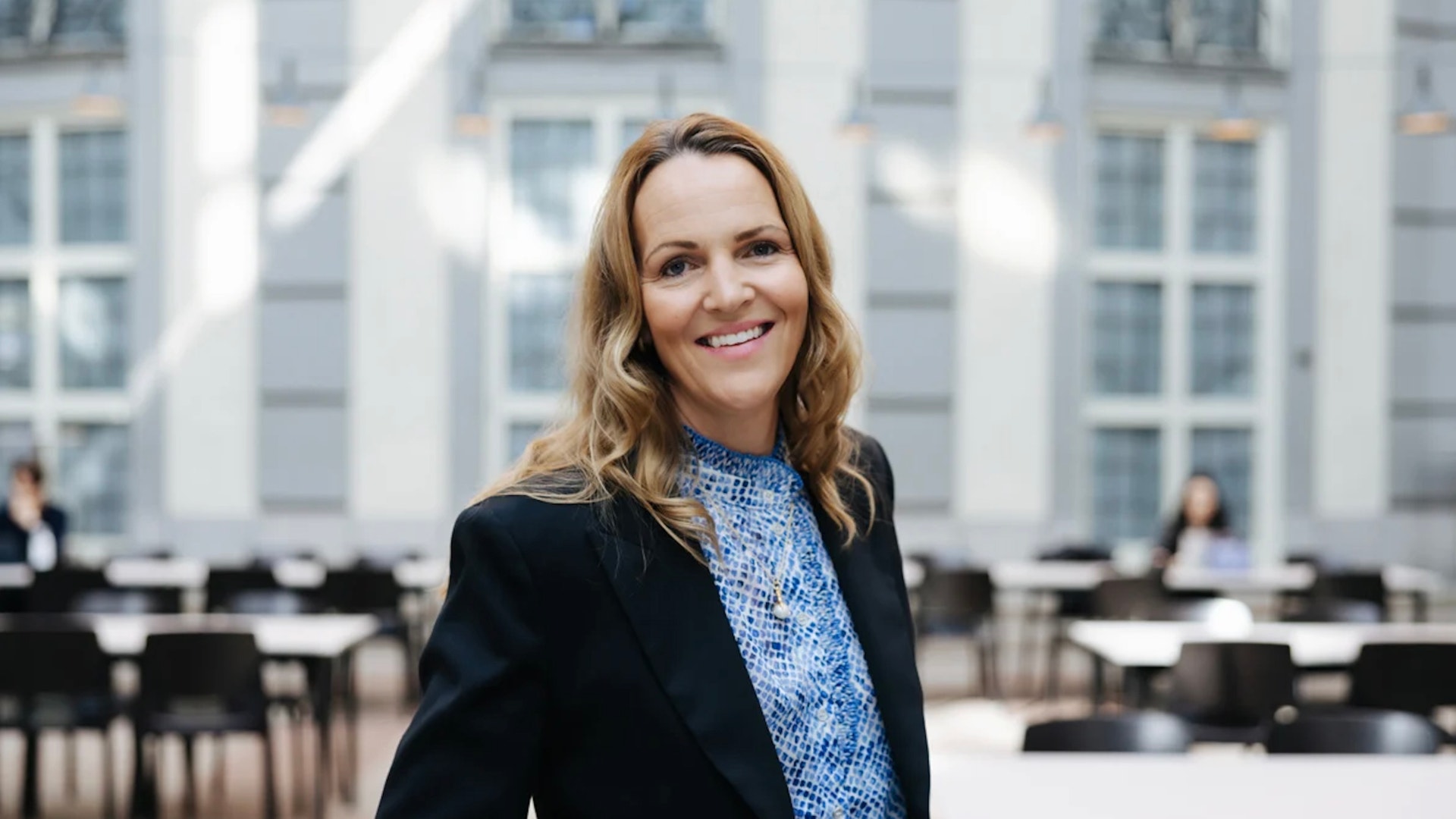 Leende kvinna med långt, vågigt hår, svart kavaj och blåmönstrad blus i en ljus lokal med bord och stolar.