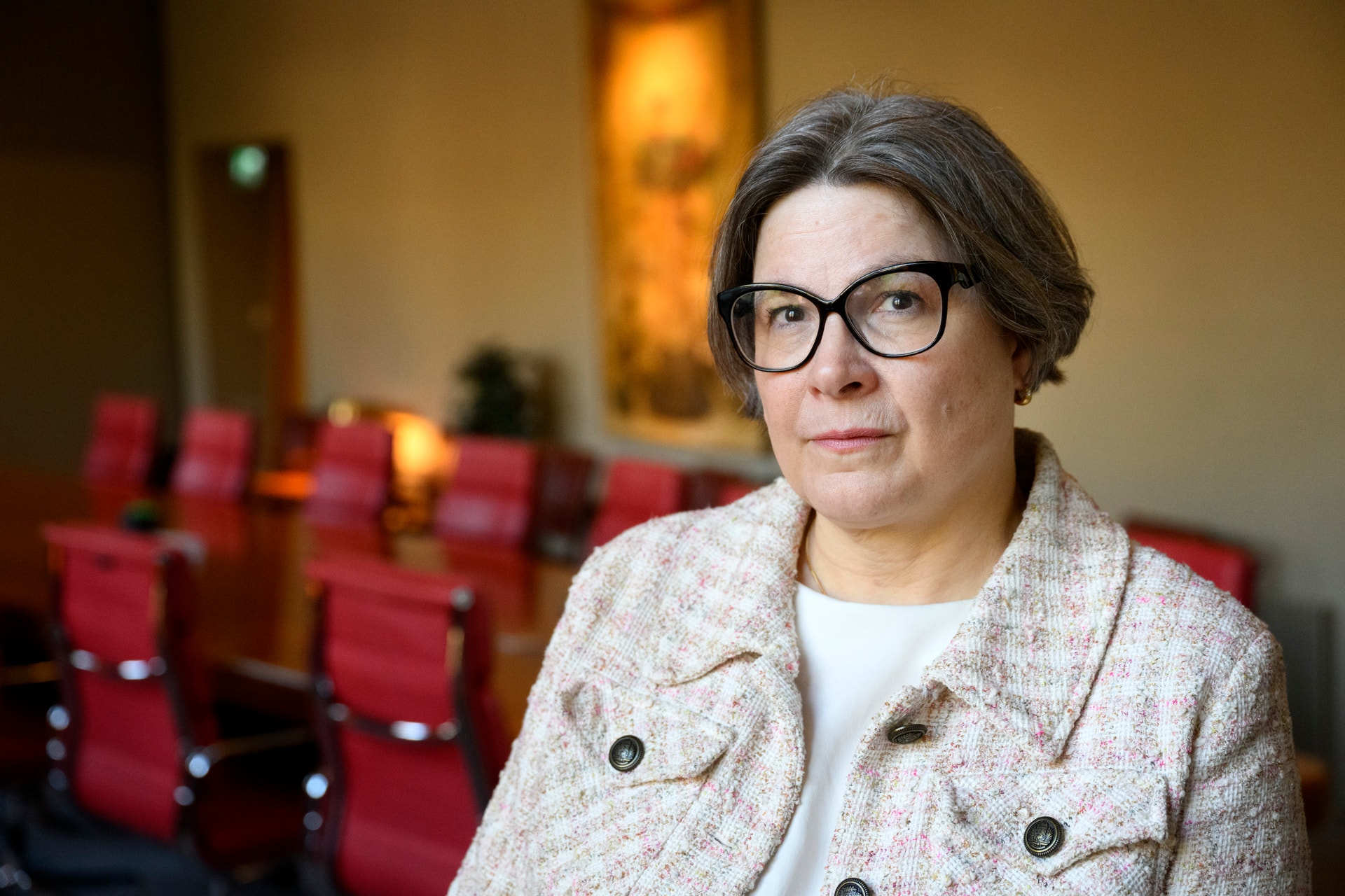 Kvinna med glasögon sitter i en auditorium med röda stolar i bakgrunden.