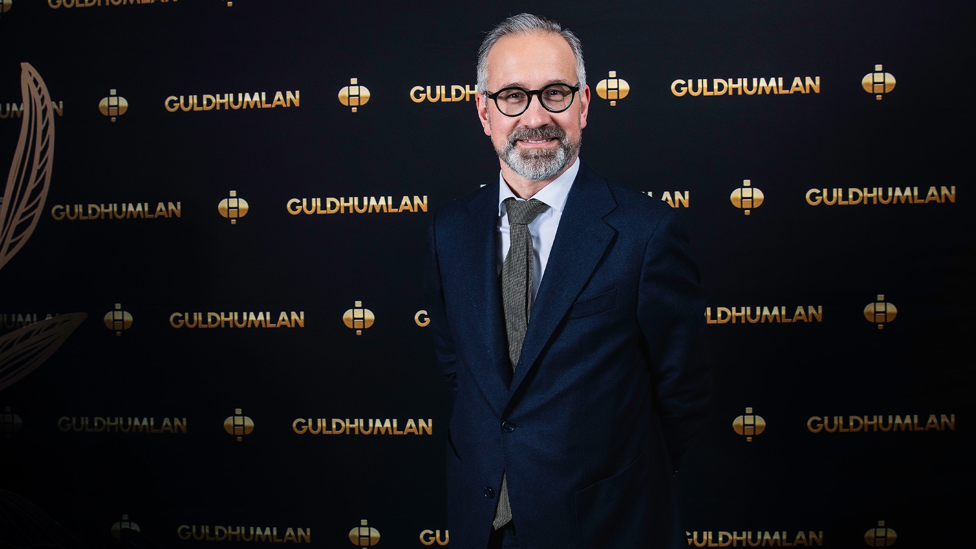 Porträttfoto av en man i mörk kostym och glasögon framför en vägg med Guldhumlan-logotyper.