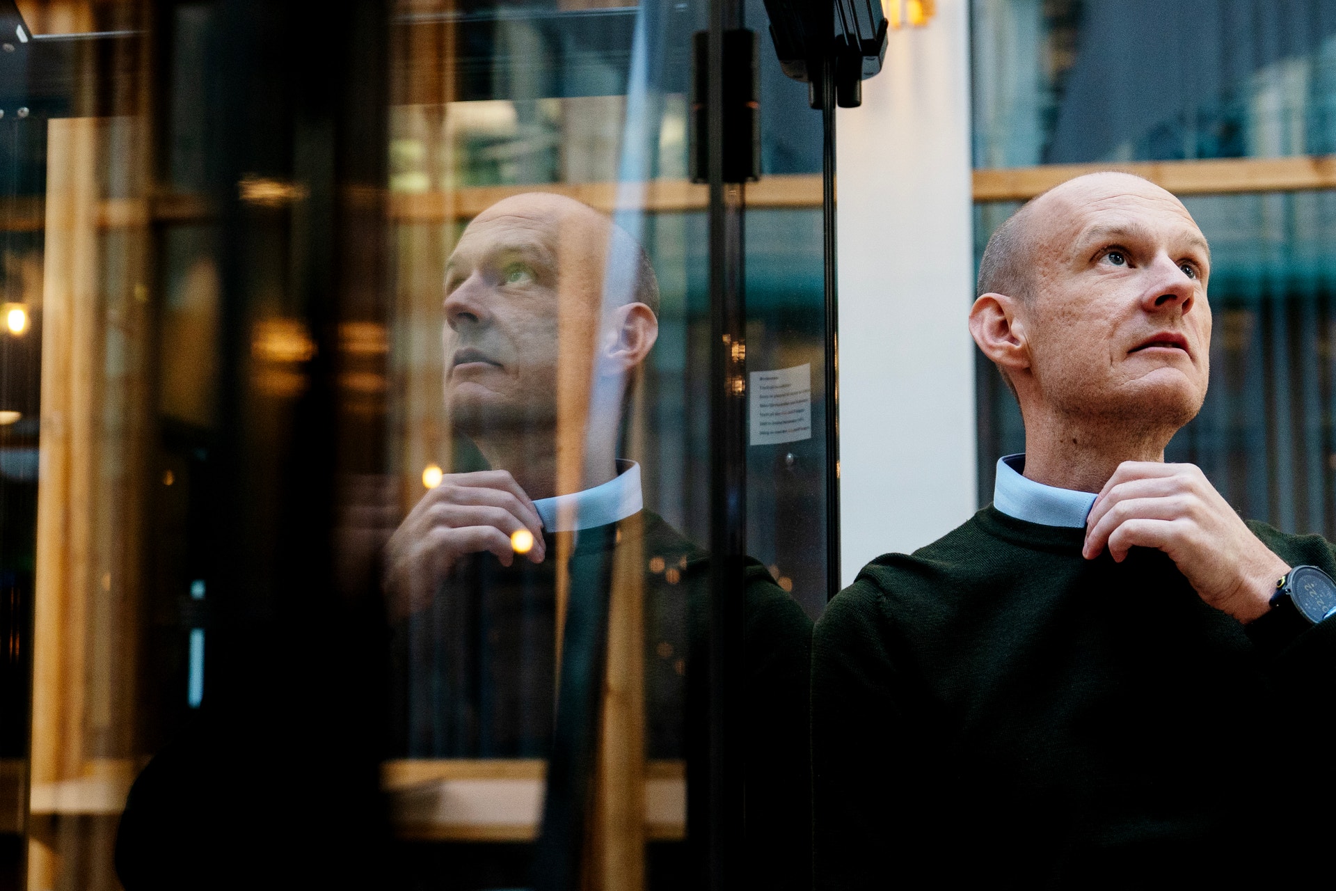 Man med kal huvud och mörk tröja tittar uppåt framför en glasdörr där han speglas.