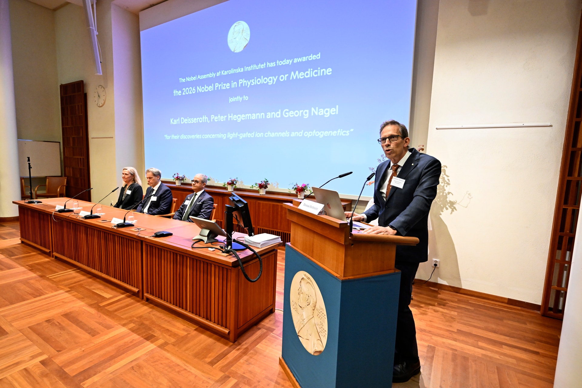 Presskonferens där Nobelförsamlingen tillkännager Nobelpriset i fysiologi eller medicin. En talare står vid en talarstol med Nobelmedaljongen, bakom honom sitter en panel med fyra personer framför en skärm med information om pristagarna.