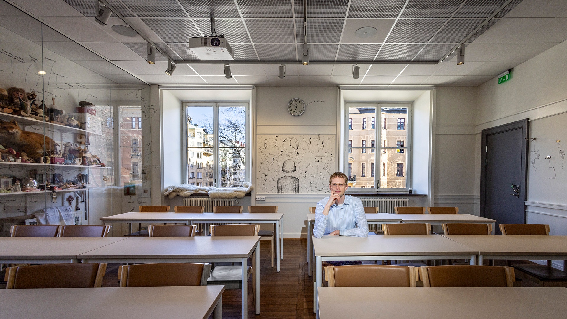 En man sitter vid ett skrivbord i ett ljust klassrum med stora fönster och träbänkar.