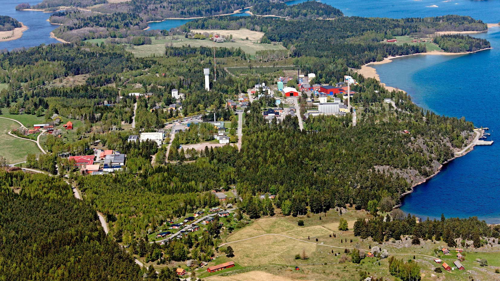 Flygfoto över Studsvik med industribyggnader och anläggningar omgivna av grön skog och blå vatten.