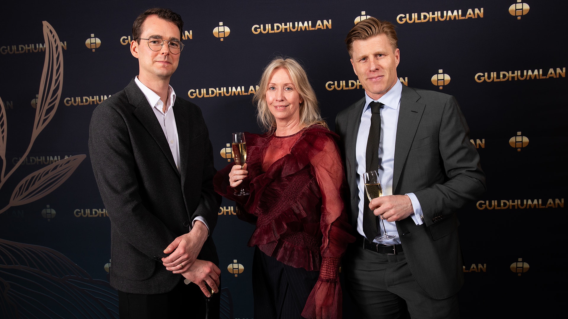 Tre personer i formell klädsel står framför en vägg med Guldhumlan-logotyper. Två av dem håller champagneglasögon.