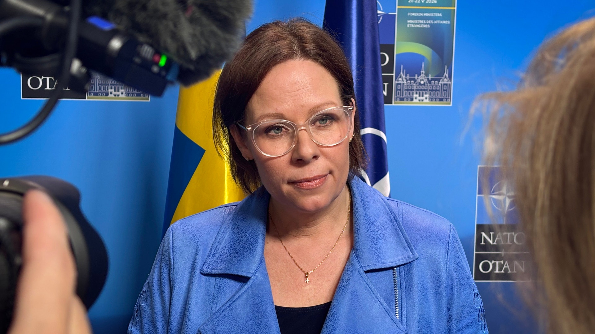 Porträttbild av en kvinna med glasögon och ljusblå skjorta framför en vägg med svenska flaggan och NATO-logotyper.
